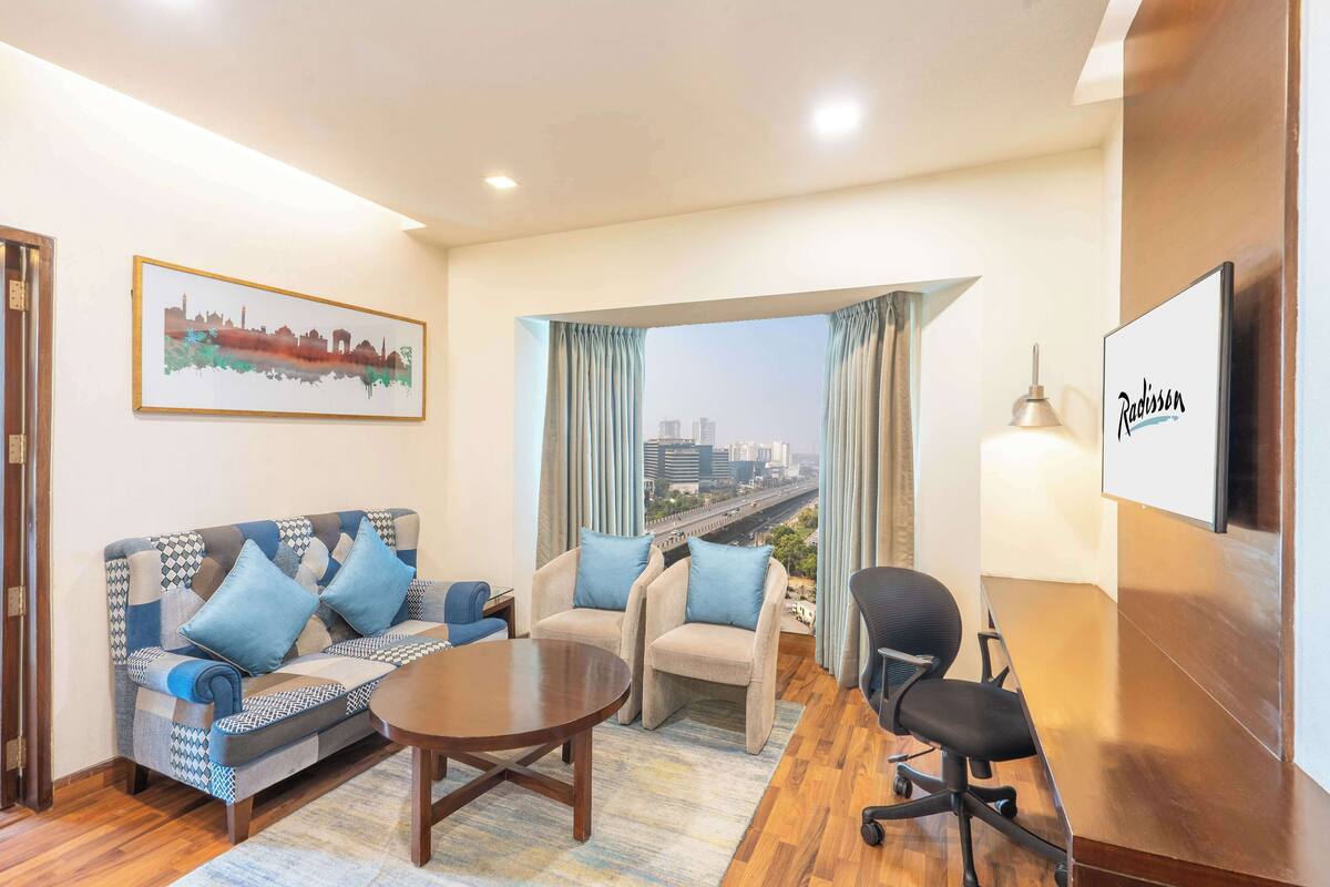 Quarto de hotel moderno com vista para a cidade, um sofá, duas poltronas, uma mesa de jantar e uma televisão.
