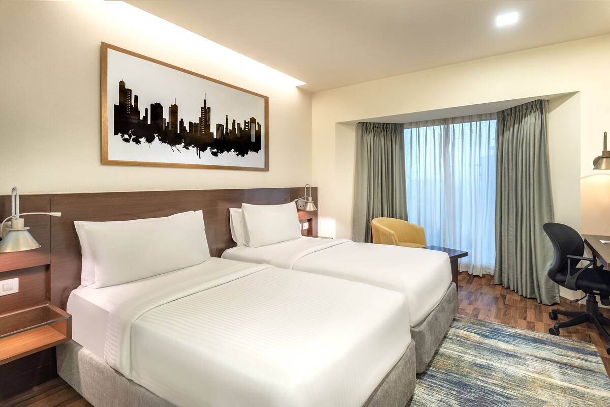 Quarto de hotel com duas camas, uma escrivaninha, uma cadeira, luminária de mesa, uma obra de arte com paisagem urbana e uma janela com cortinas.