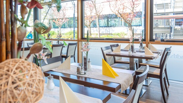 A restaurant interior with tables set for dining, large windows, and indoor plants. - Hotel an der Stadthalle Rostock-Hbf (Rostock)