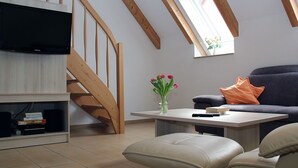 A modern loft-style living room with a wooden staircase, a flat-screen TV, a coffee table, a sofa, and a vase with flowers.