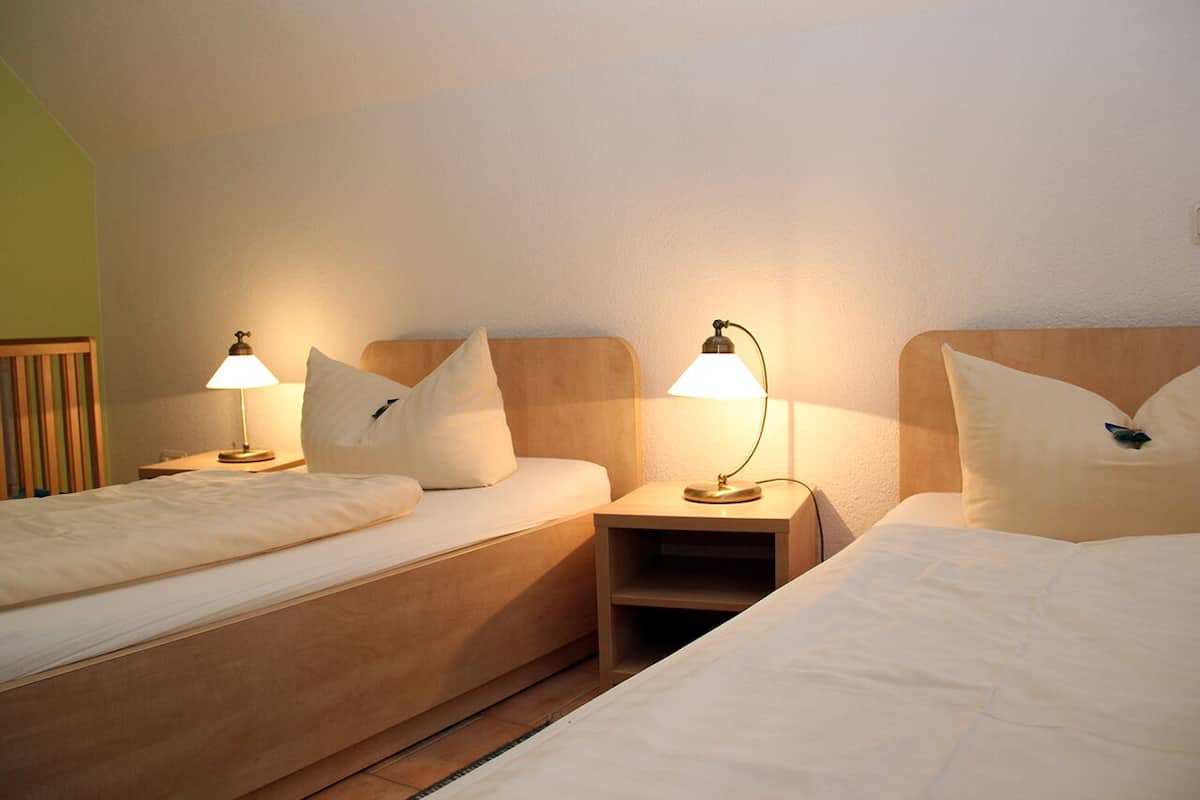 Quarto de hotel com duas camas, cada uma com cabeceira de madeira, mesa de cabeceira e abajur.