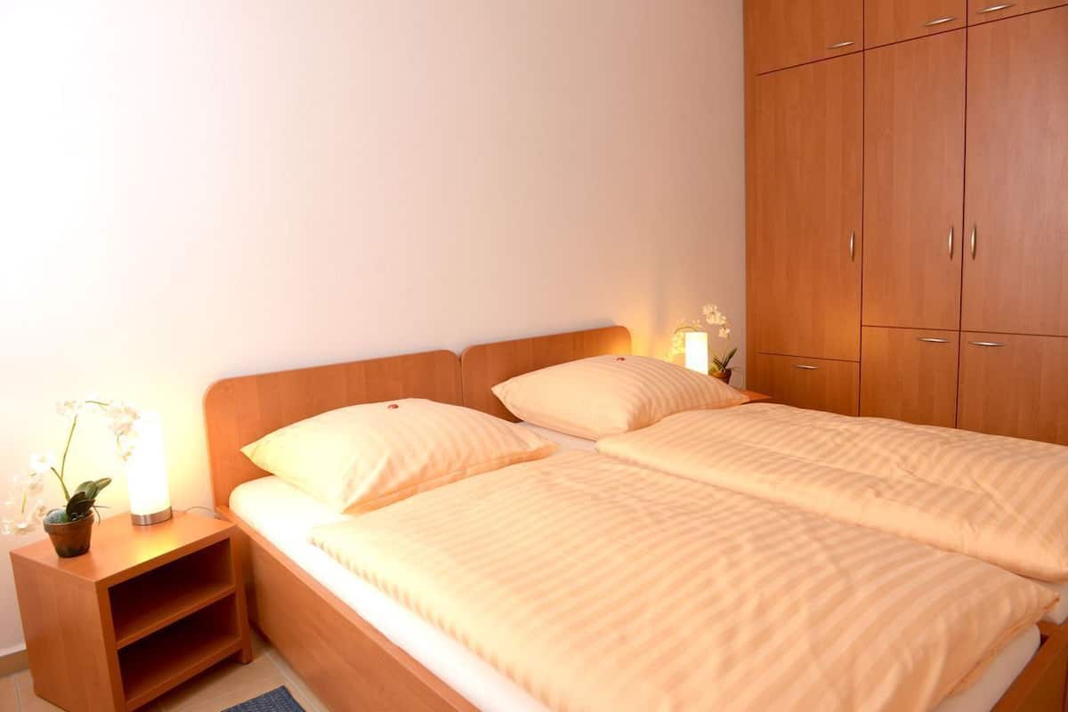 Uma cama de casal com duas almofadas, um criado-mudo com abajur e planta em vaso, e um guarda-roupa de madeira.