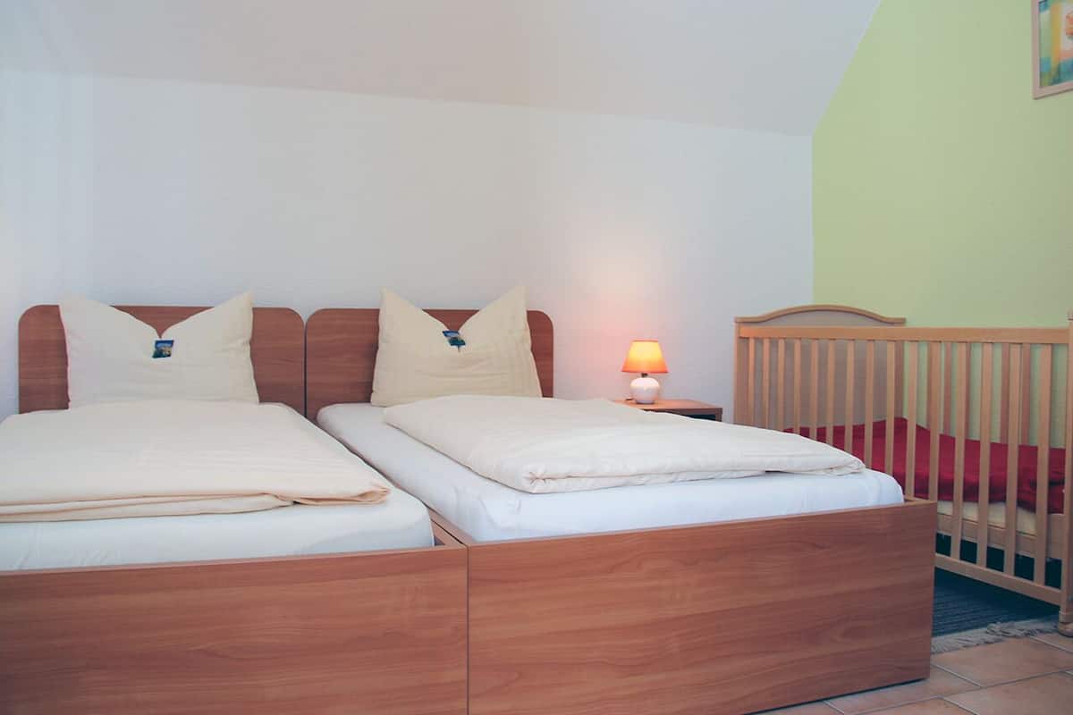 Uma cama de casal com estrutura de madeira, uma mesa de cabeceira com abajur e um berço em um quarto com paredes verde-claras.