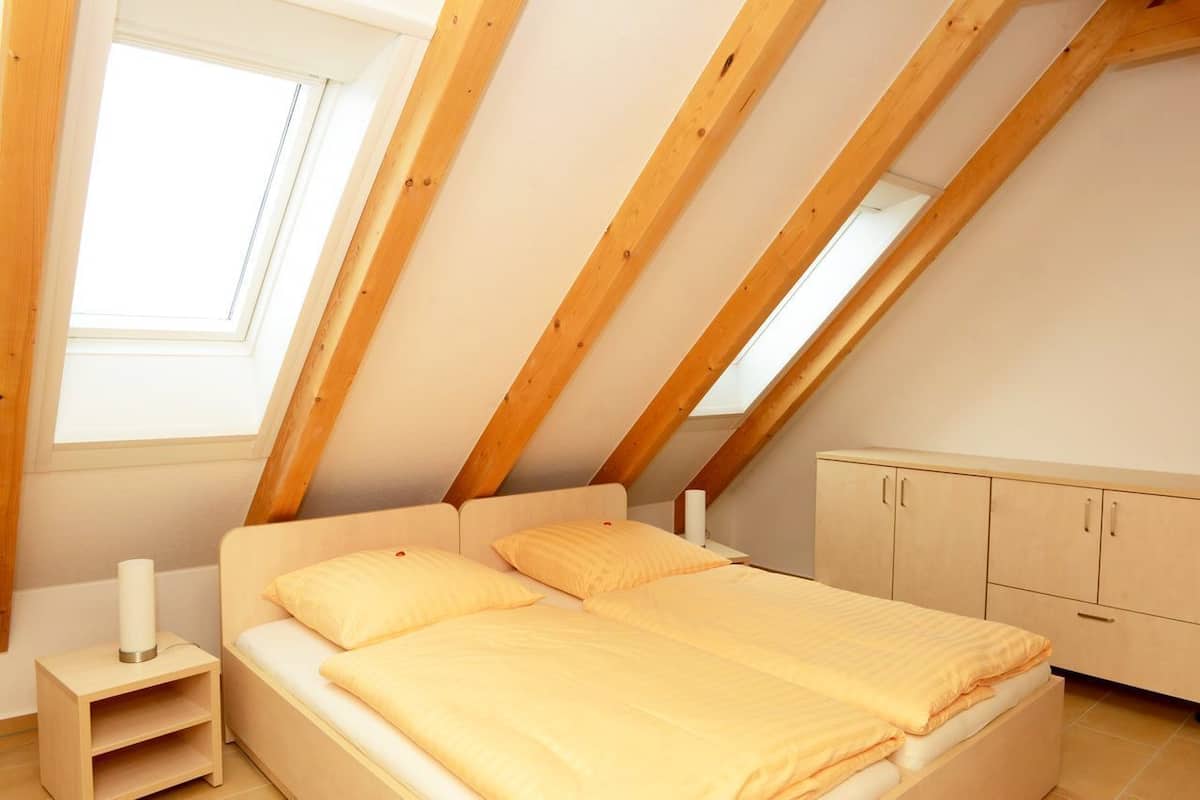 Quarto com teto inclinado, uma cama com duas almofadas, uma mesinha de cabeceira e um guarda-roupa.