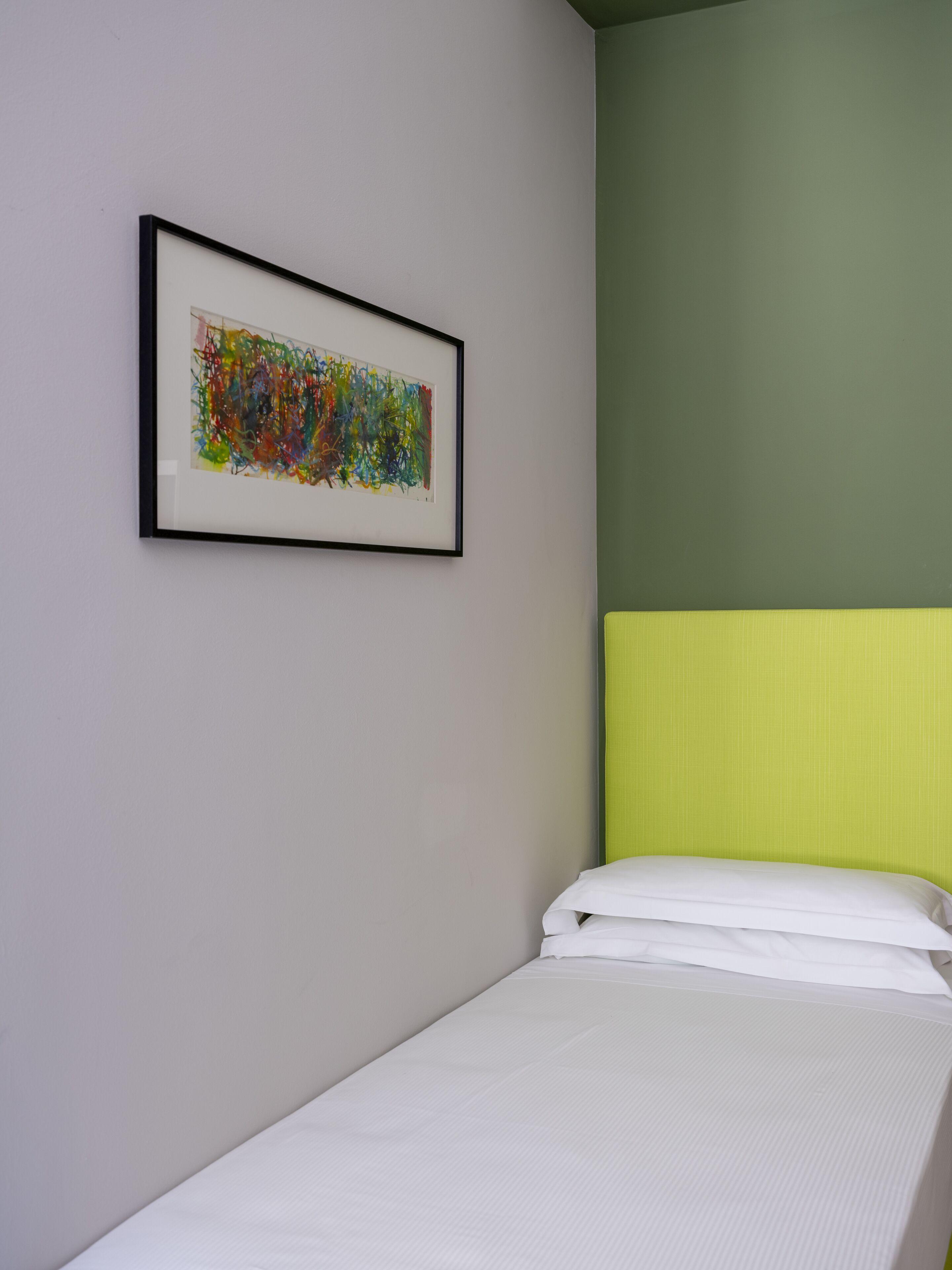 A single bed with white bedding in a room with a green wall and a framed abstract painting.