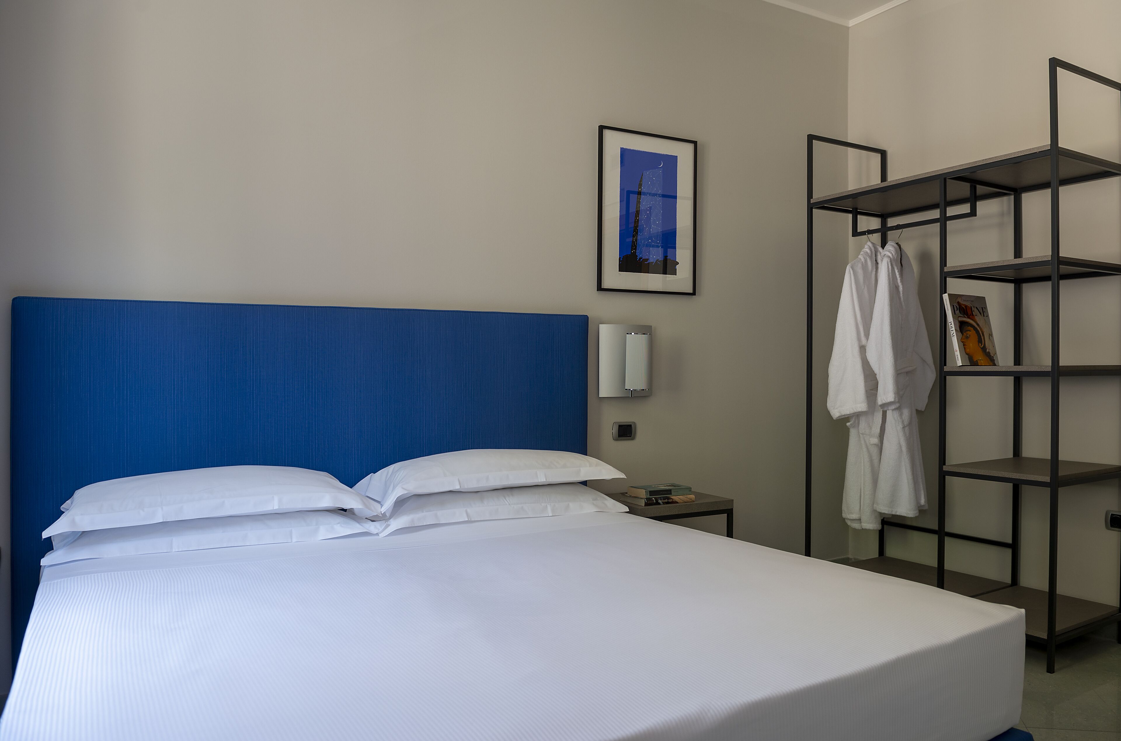 Habitación de hotel con una cama grande, cabecera azul, pared blanca, un cuadro enmarcado, una estantería con ropa y una mesita de noche con lámpara.