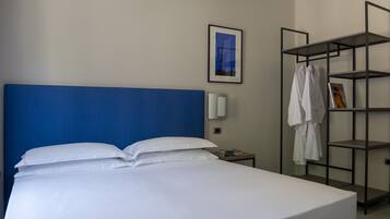 Quarto de hotel com uma cama grande, cabeceira azul, parede branca, quadro emoldurado, prateleira com roupas e mesa de cabeceira com abajur.