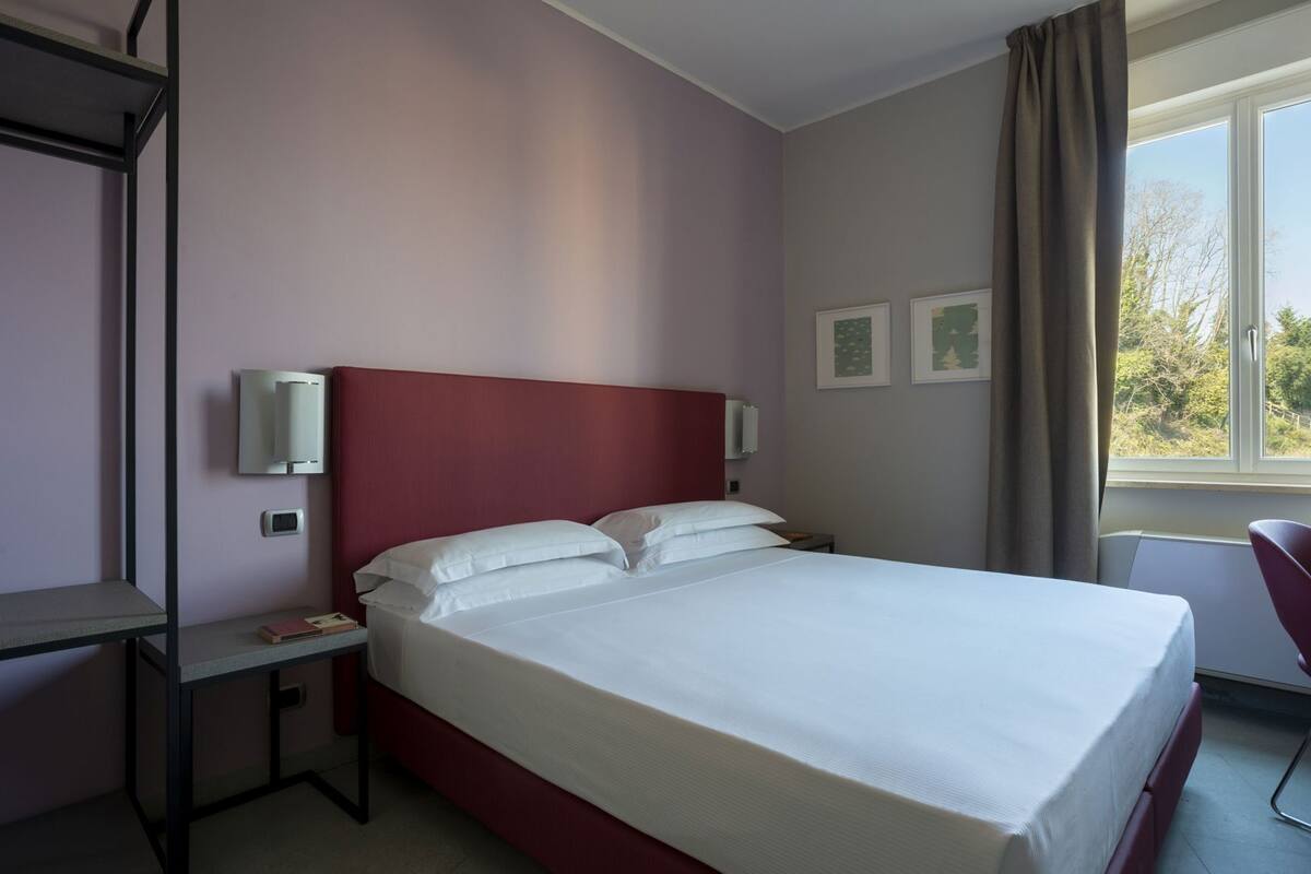Una habitación de hotel con una cama individual, mesitas de noche, un escritorio, una silla y una ventana con cortinas.