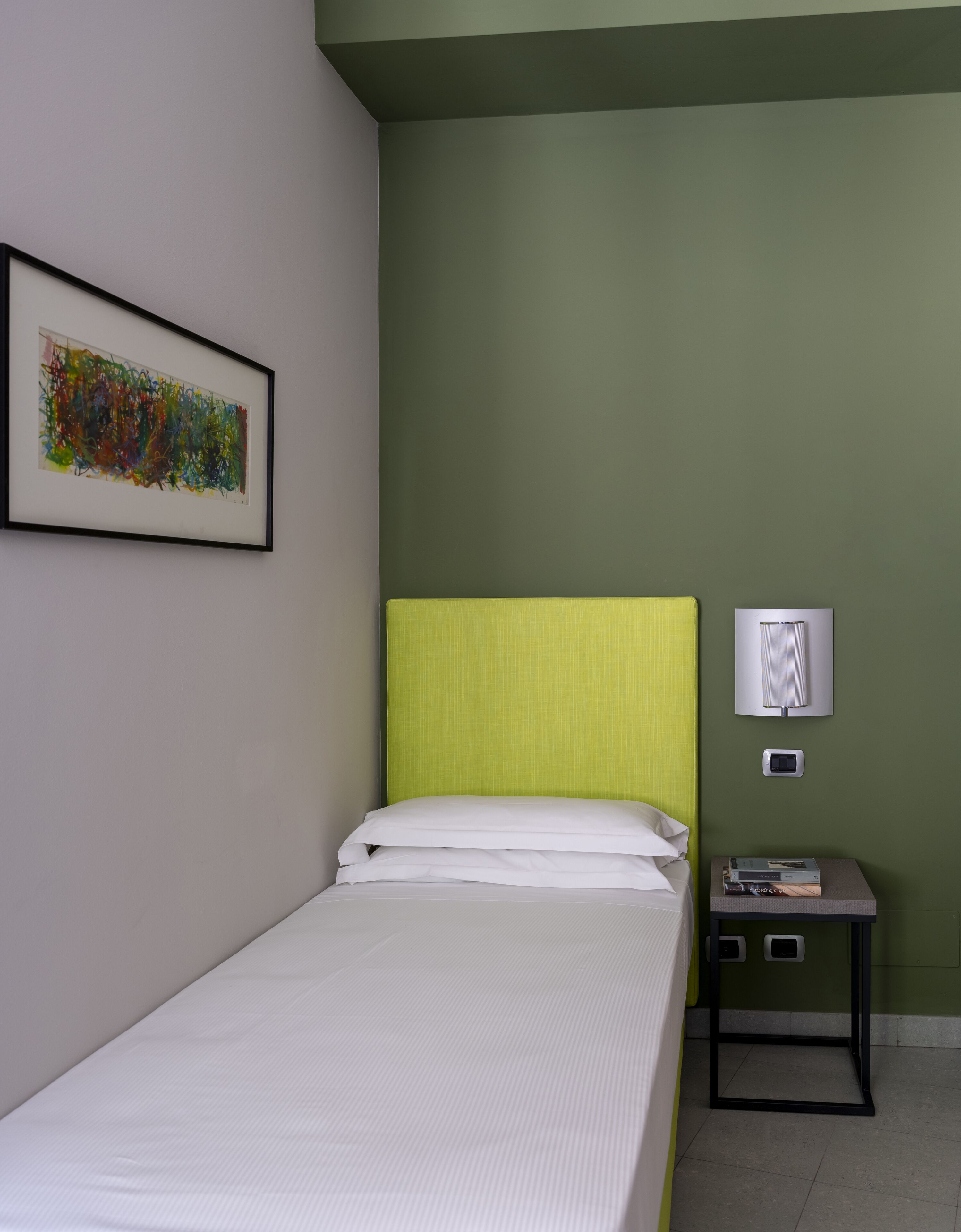 A single bed with white bedding, a green headboard, a framed abstract painting on the wall, and a small bedside table with a book and a lamp.