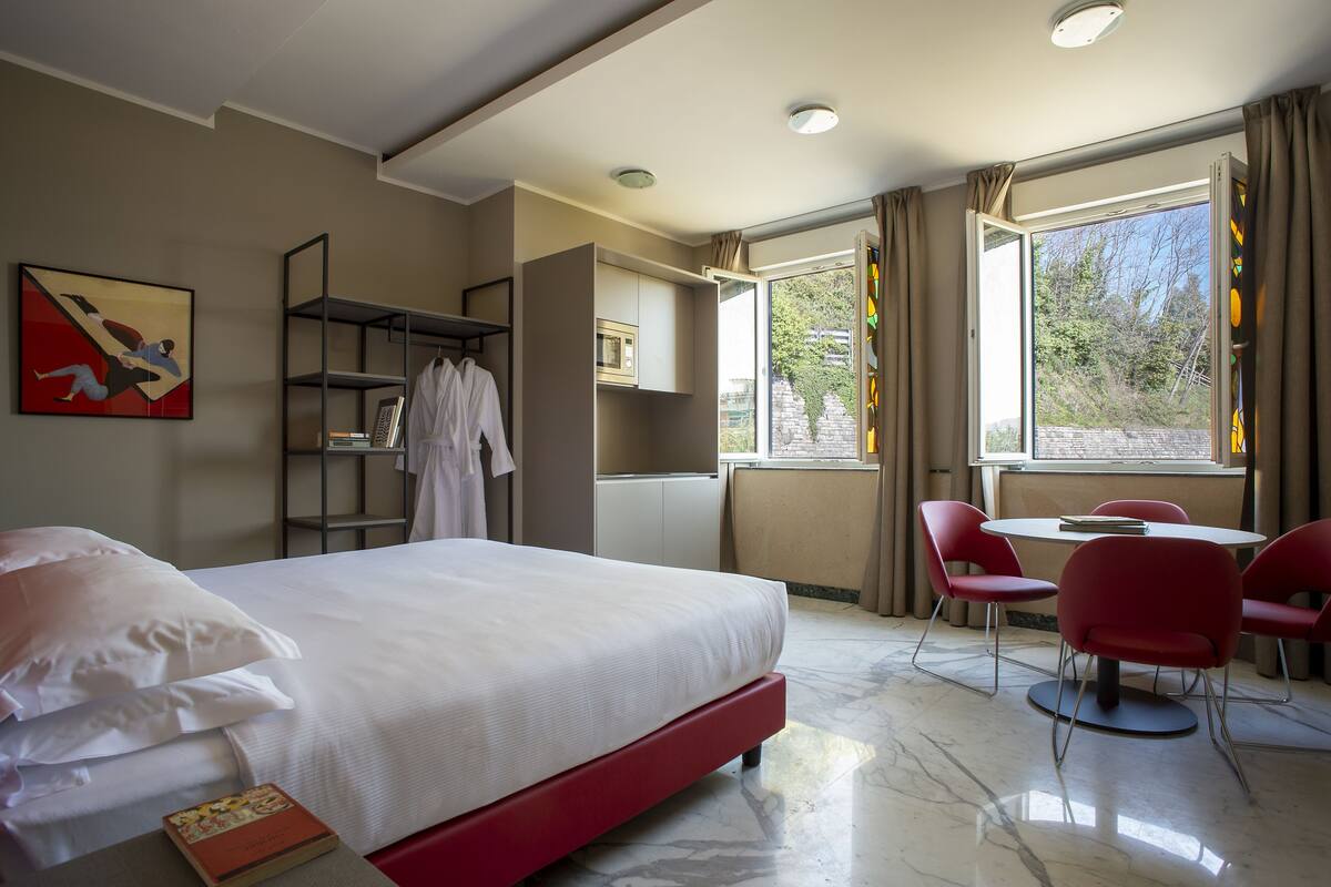 Habitación de hotel con cama, una silla roja, una mesa, una estantería y ventana con cortinas.