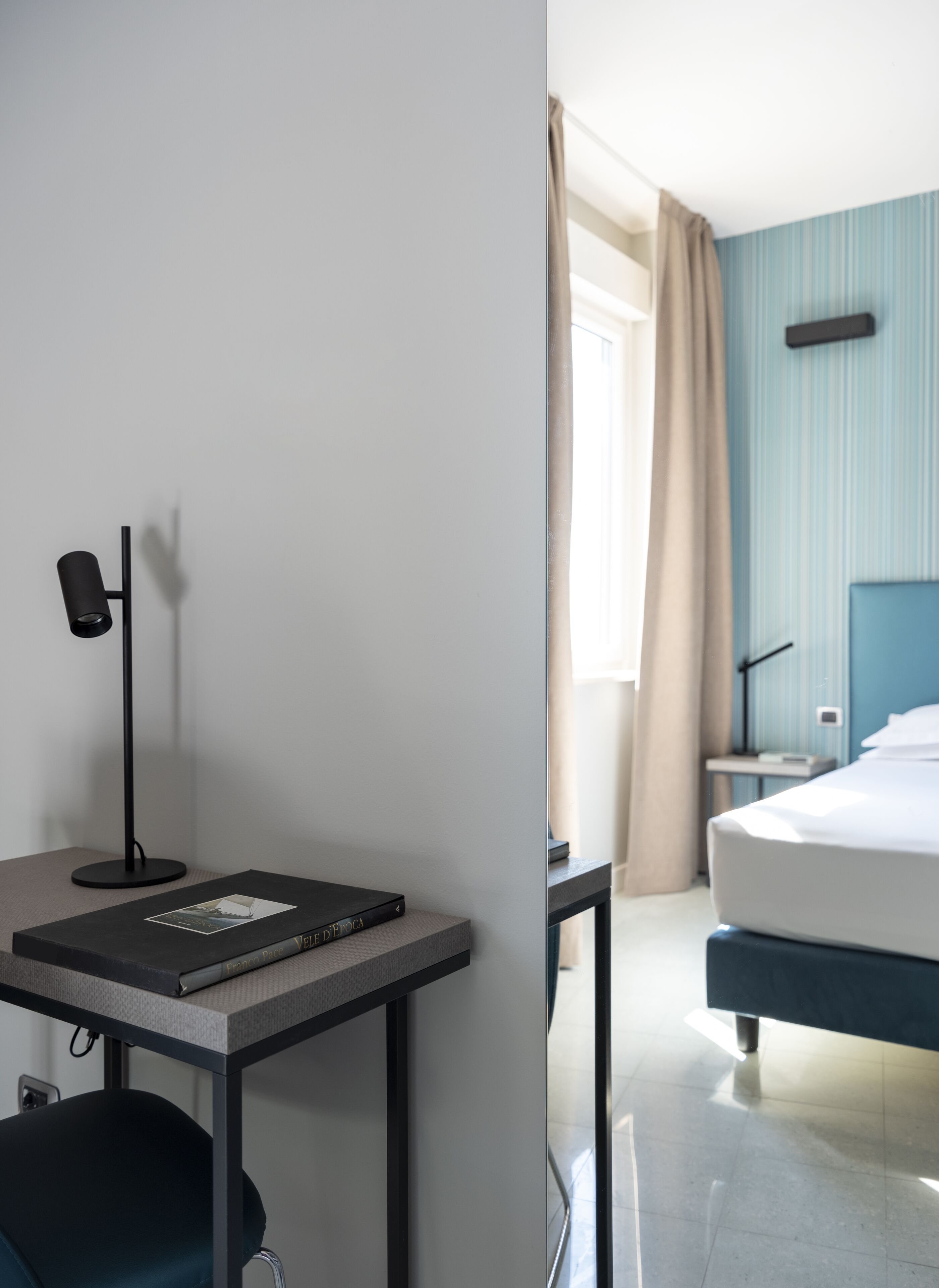 A hotel room with a bed, a desk with a lamp, a chair, a nightstand, and a window with curtains.