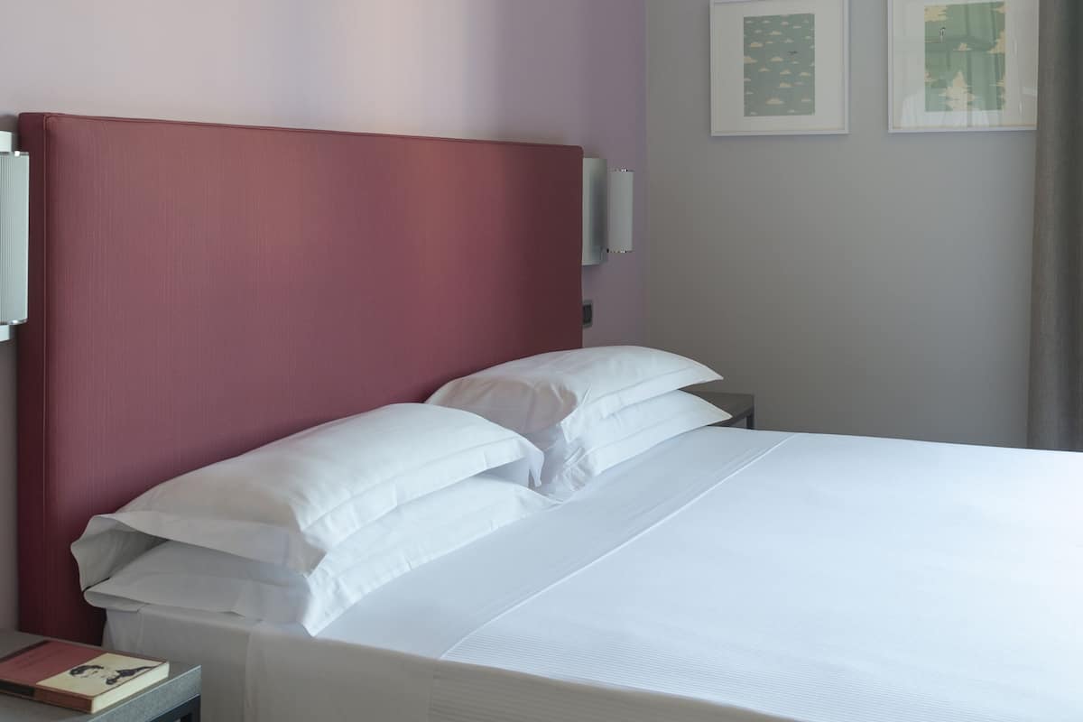 Una cama bien hecha con sábanas blancas en una habitación con cabecera roja y paredes grises claras. Hay dos cuadros en la pared y una mesita de noche con un libro.