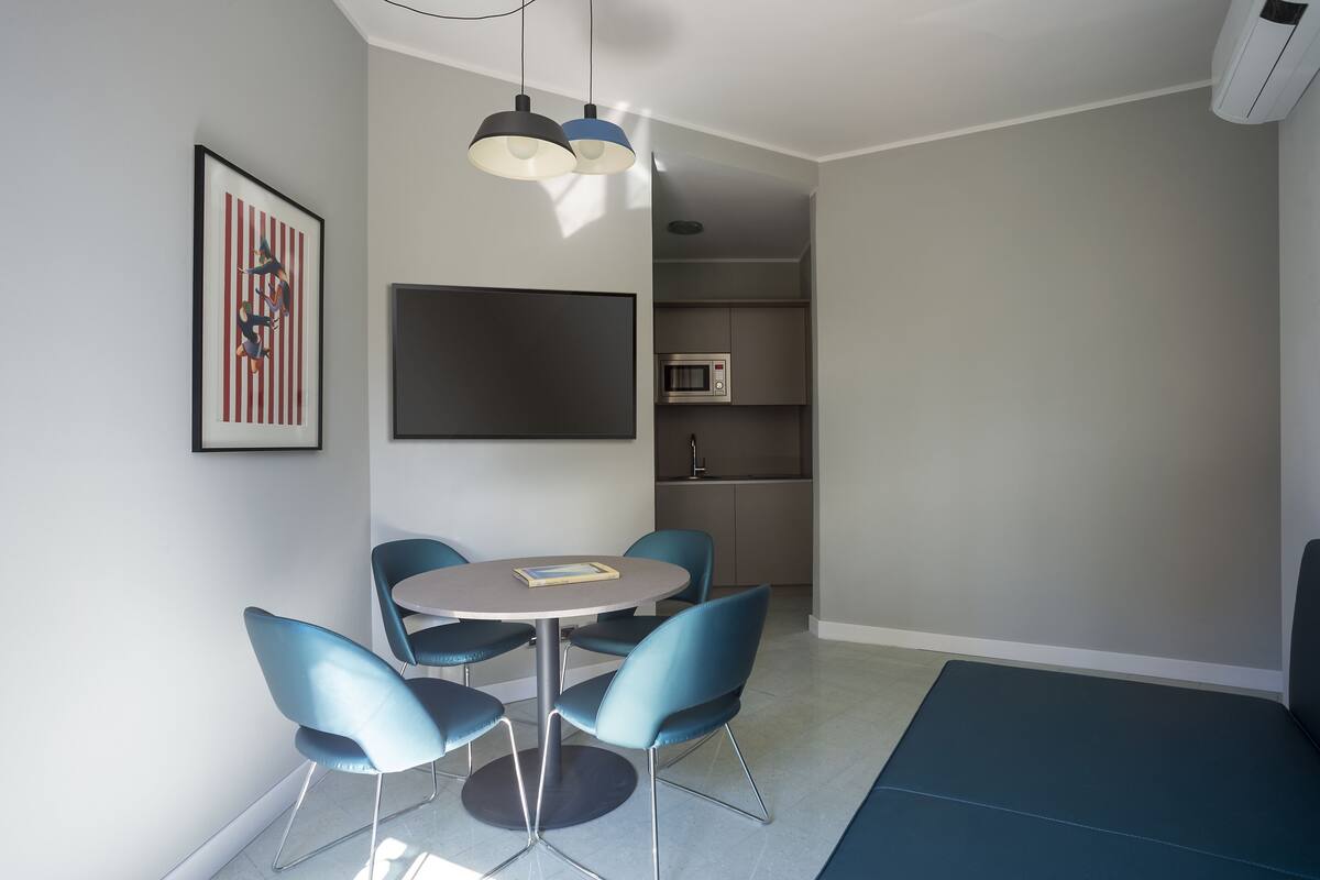 Una habitación moderna y compacta con una mesa redonda y cuatro sillas azules, un televisor de pantalla plana montado en la pared y una pequeña zona de kitchenette con microondas.