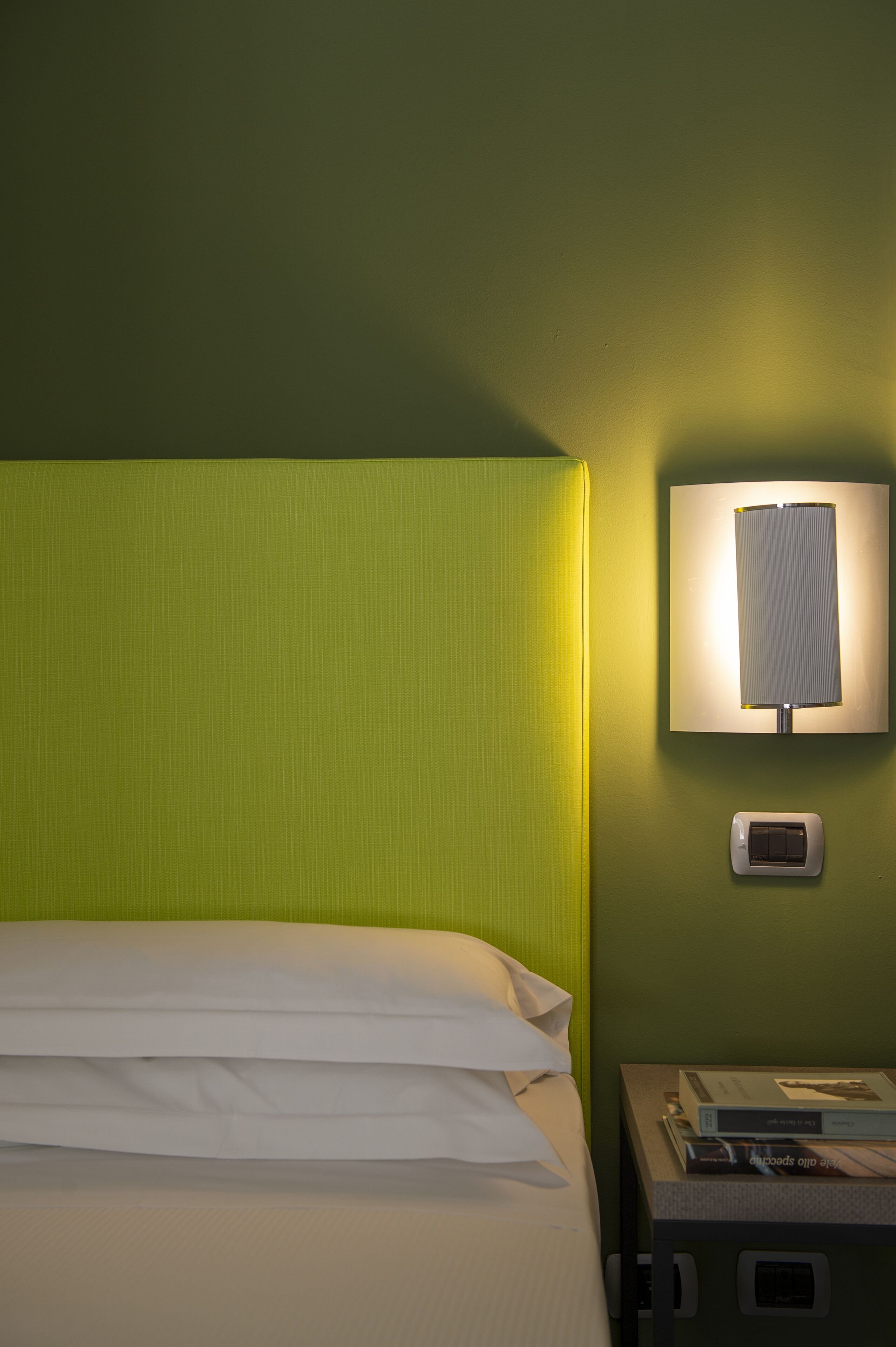 Ein Bett mit weißer Bettwäsche, eine grüne Wand, eine Wandleuchte und ein kleiner Tisch mit Fernbedienung und Büchern.