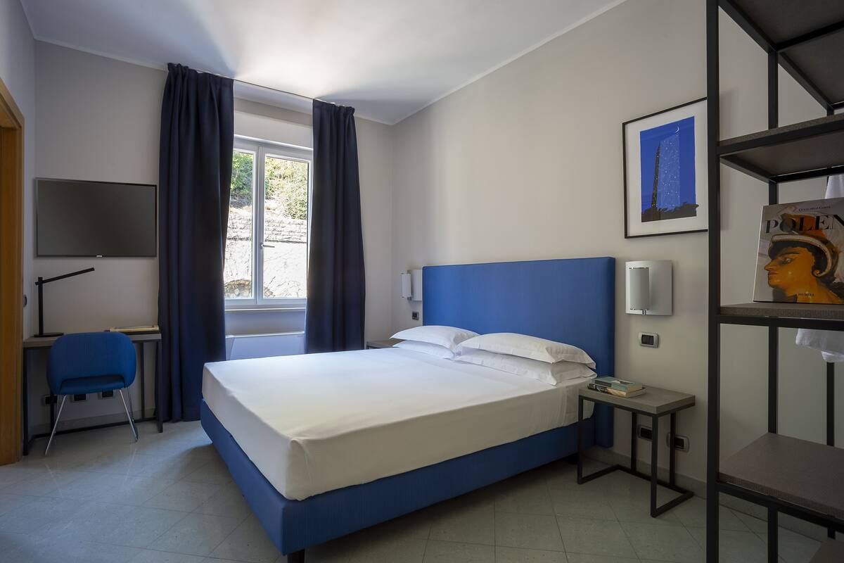 Habitación de hotel con cama, escritorio, silla, televisión, ventana con cortinas, un cuadro enmarcado y una estantería con un libro.