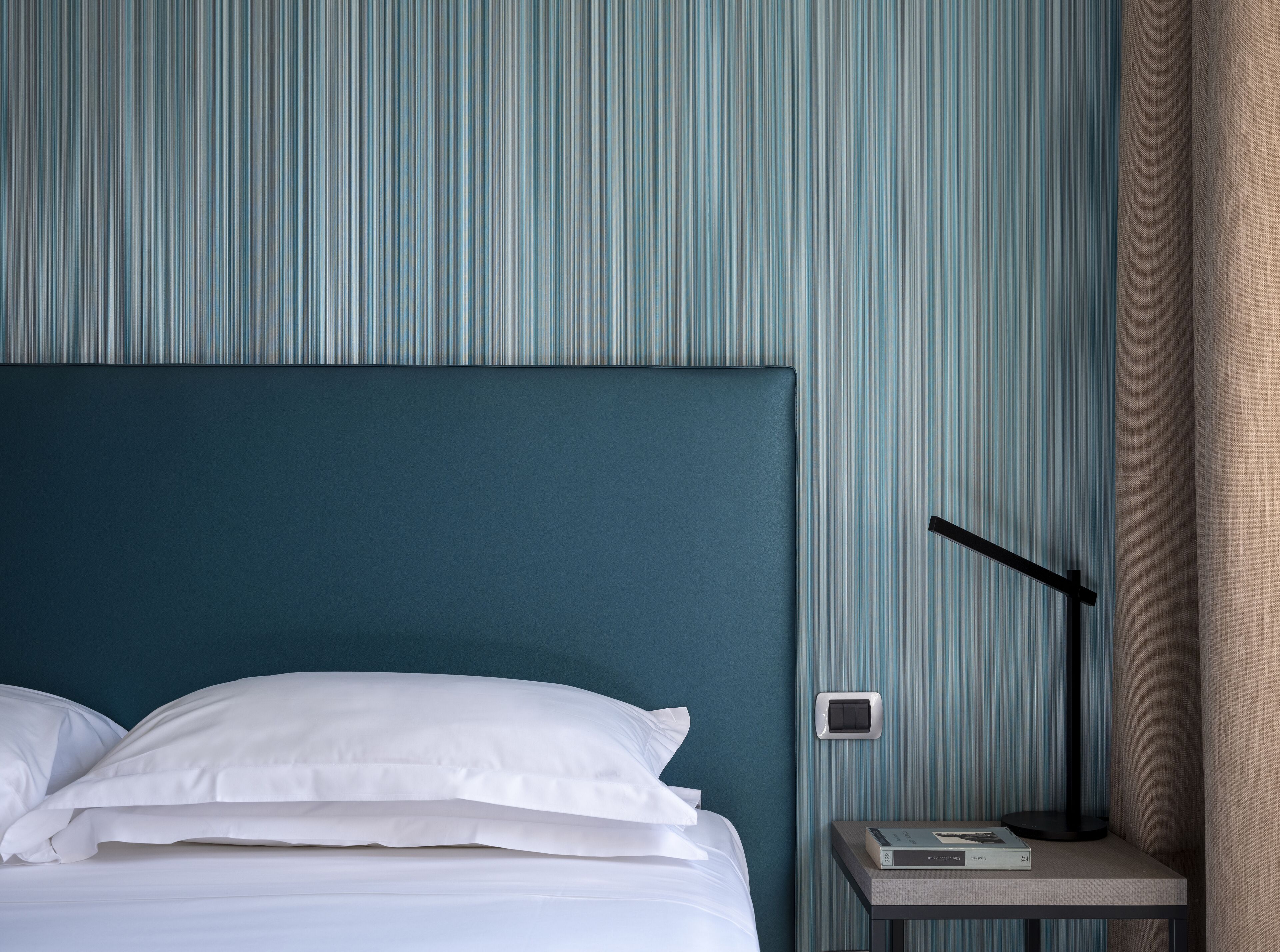 A neatly made bed with white linens, a dark blue headboard, and a bedside table with a lamp and a book.