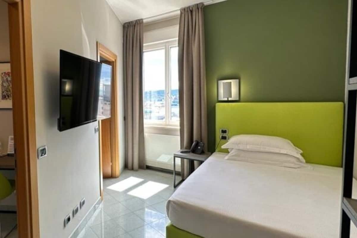 Habitación de hotel con cama, escritorio, silla, televisor y ventana con cortinas.