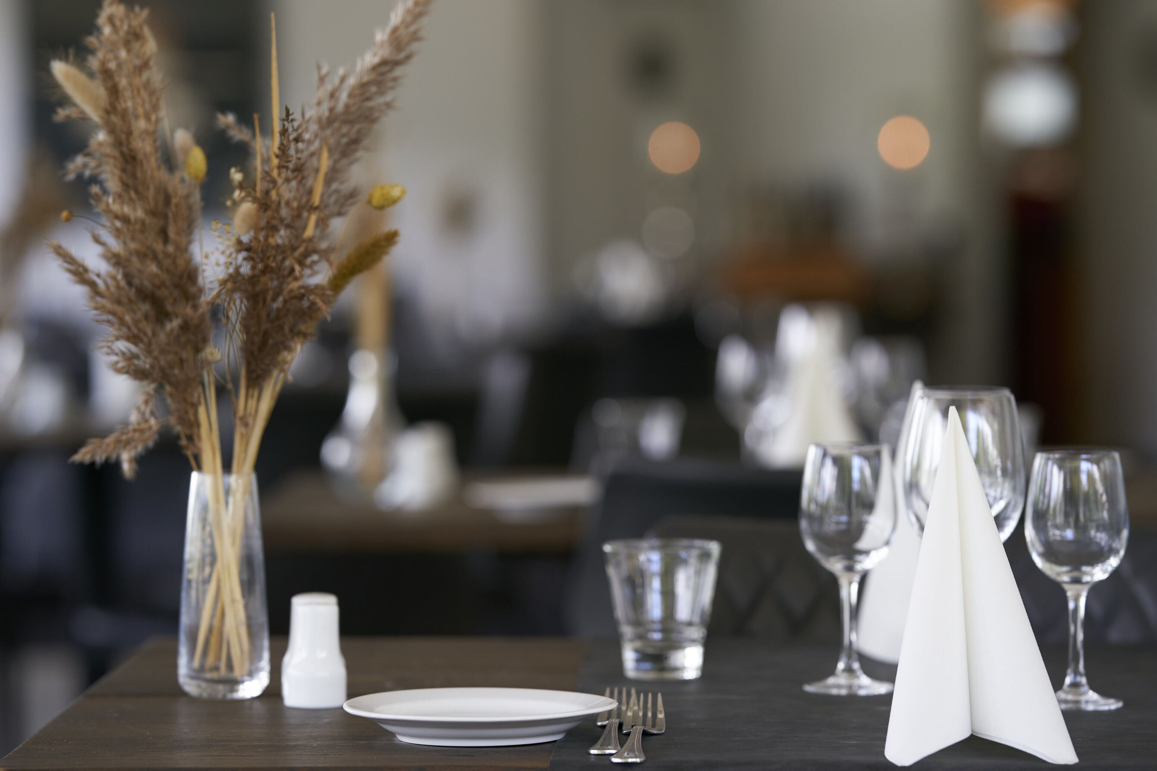 A dining table set with a white plate, silverware, a folded white napkin, and a clear glass. A vase with dried pampas grass is also visible.