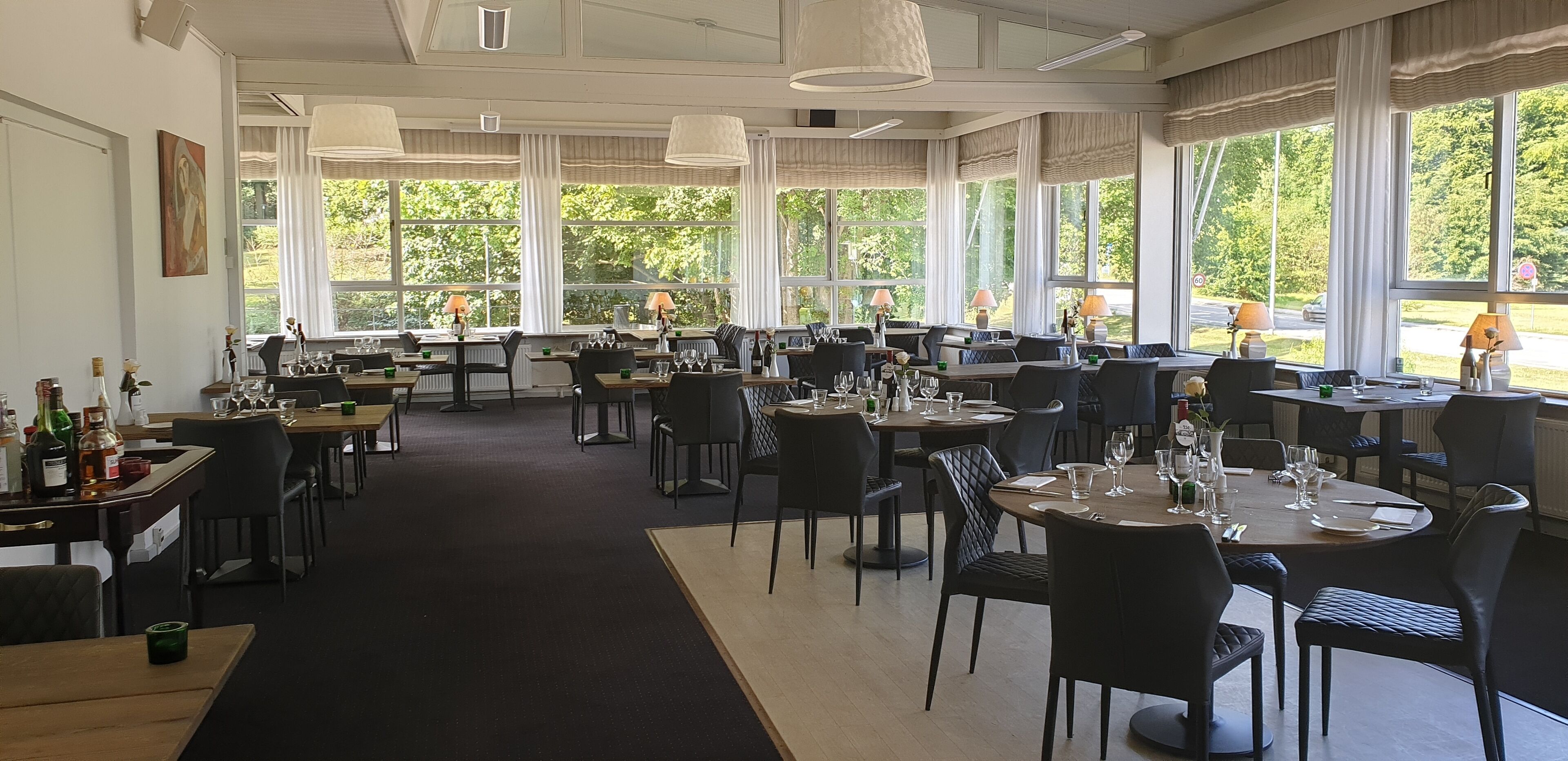 En rummelig restaurant med flere spiseborde og stole, store vinduer med udsigt til det fri, og et rent, moderne interiør.