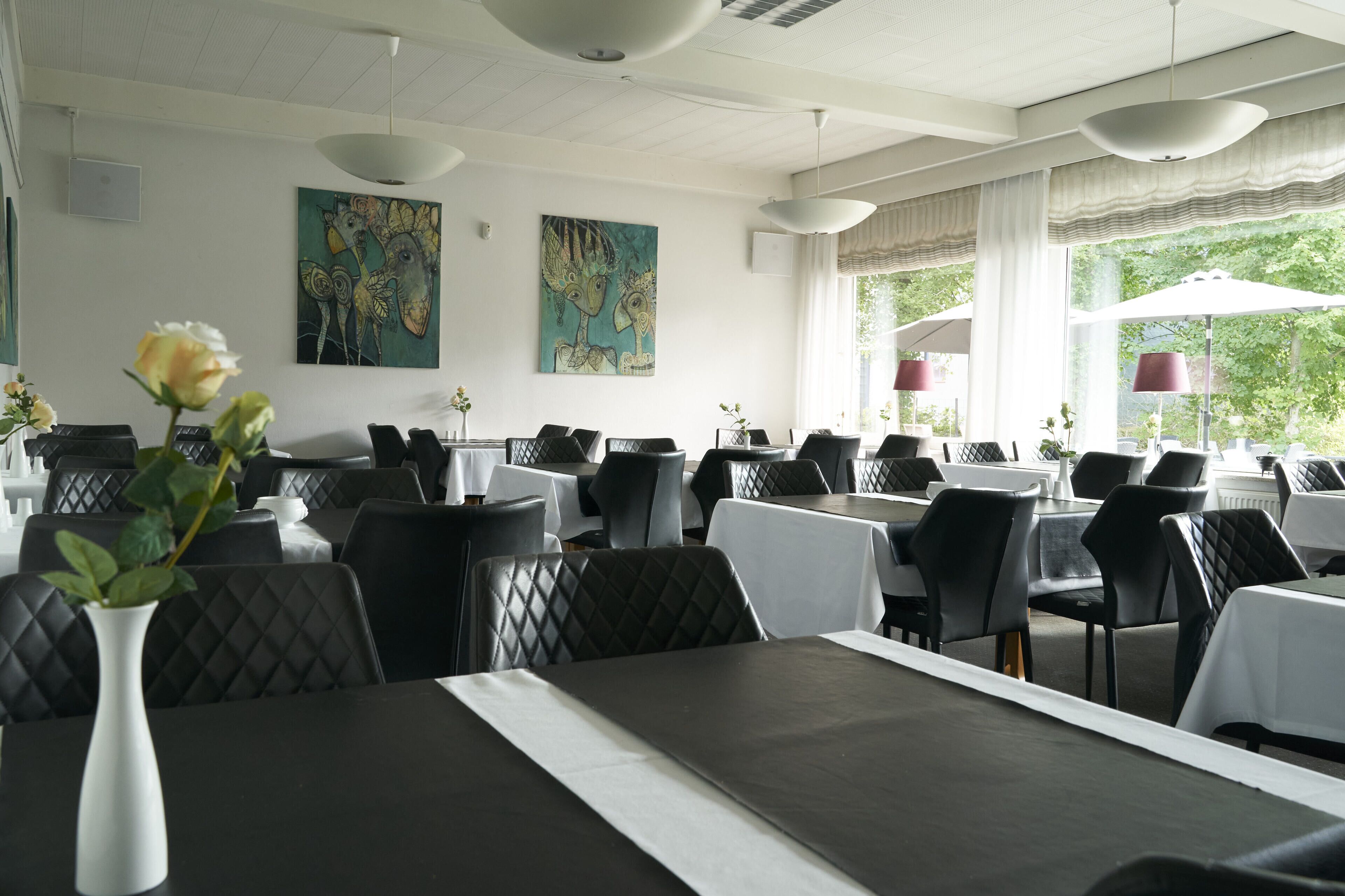 Restaurante com mesas preparadas para refeições, cadeiras estofadas pretas e obras de arte nas paredes.