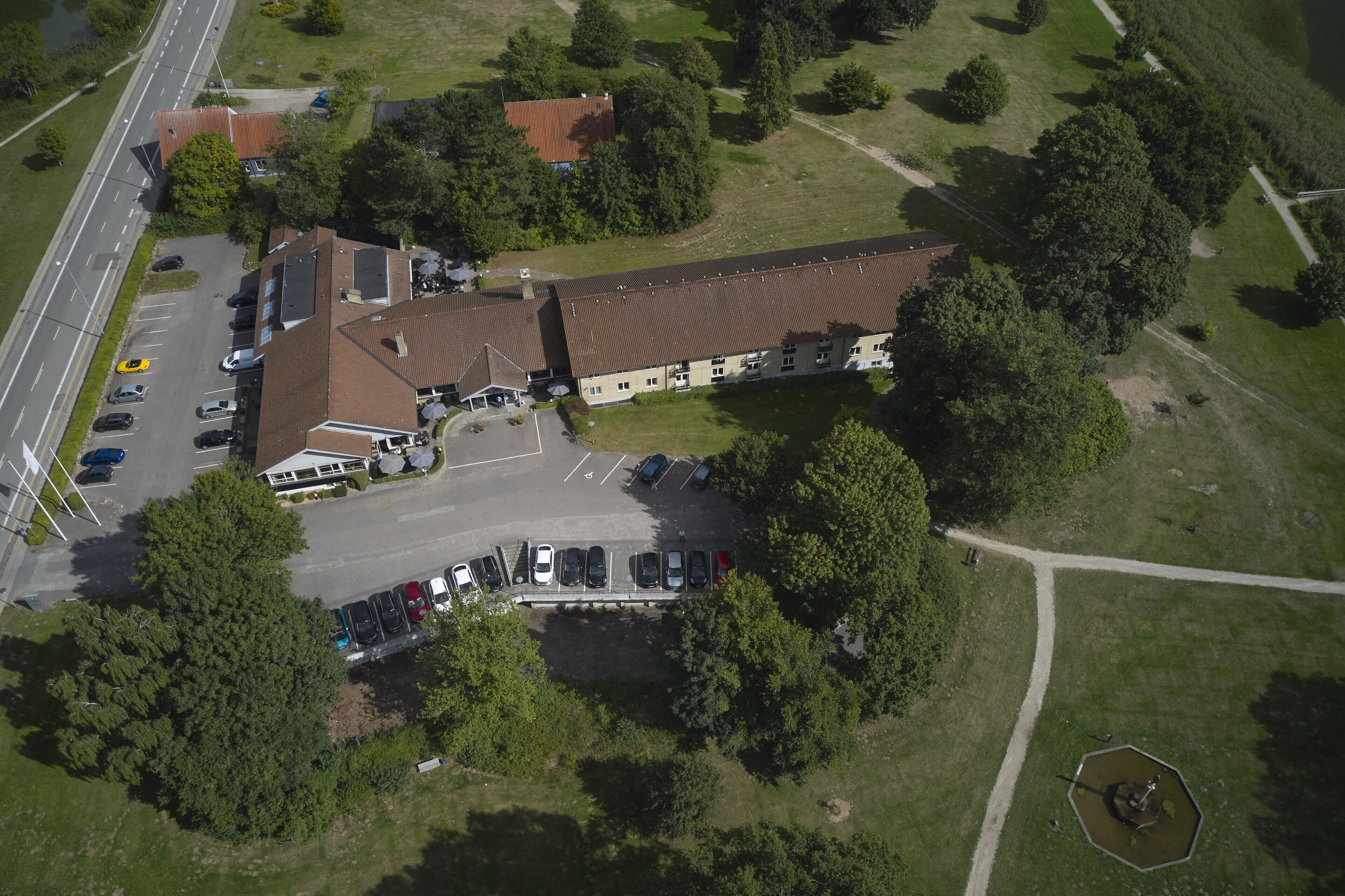 An aerial view of a building with a parking lot, surrounded by trees and a road.