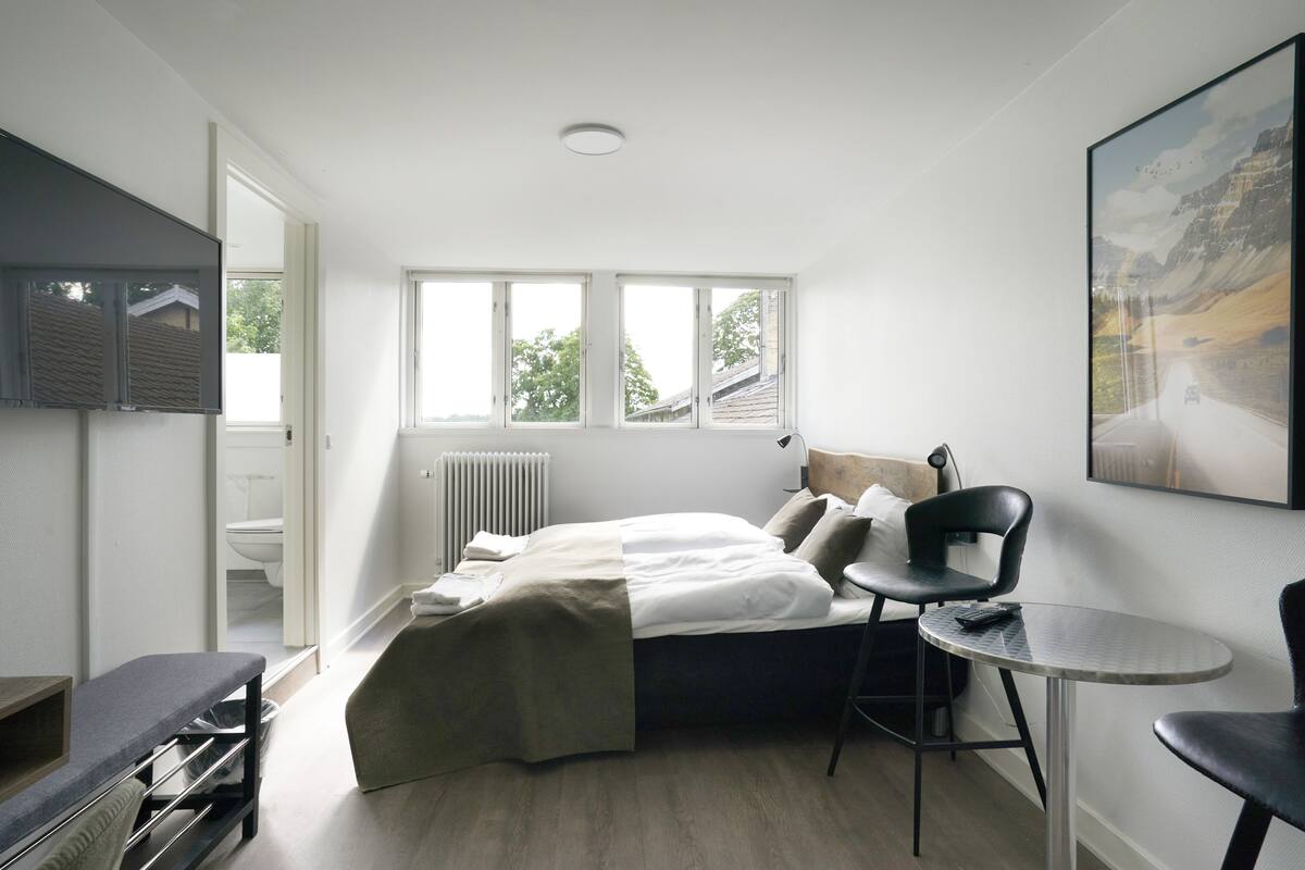 A modern bedroom with a bed, a chair, a small table, and a large window.