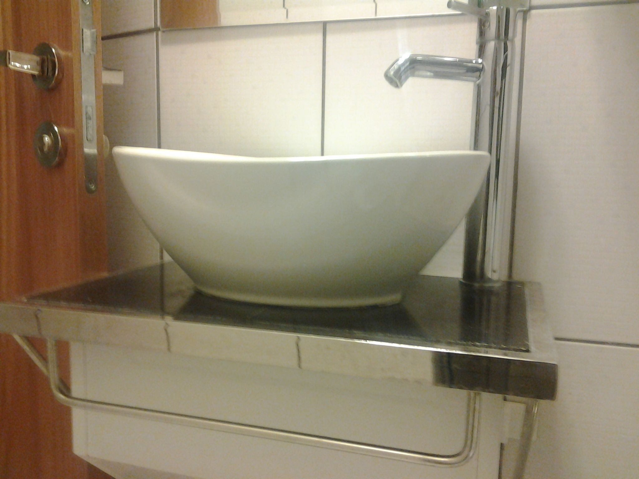 Un lavamanos blanco sobre una encimera de acero inoxidable en una cocina con baldosas blancas y una puerta de madera.