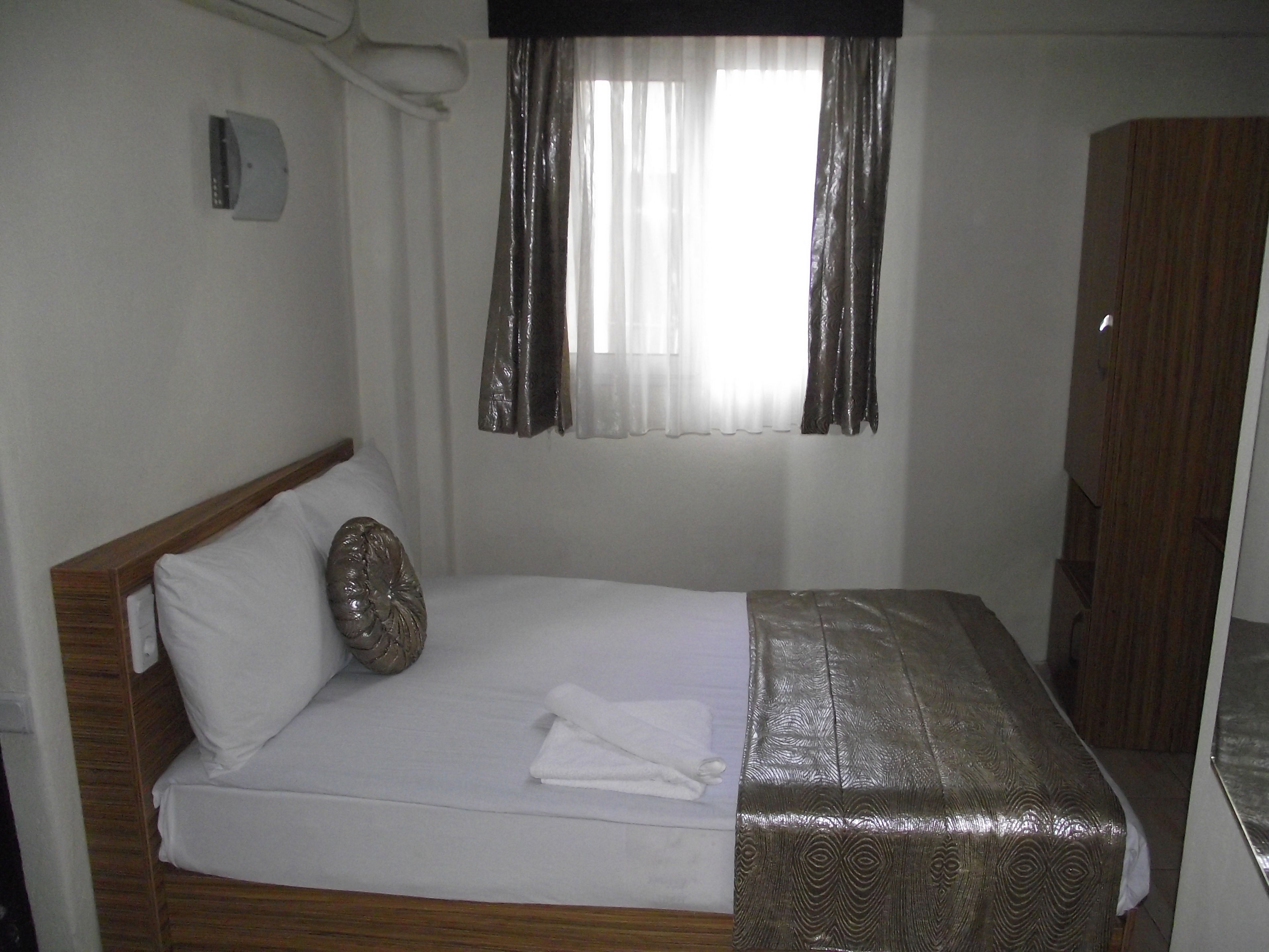 Un lit simple avec une tête de lit en bois, un coussin décoratif et une couvre-lit à motifs. Il y a une fenêtre avec des rideaux légers et un rideau plus épais, ainsi qu’une armoire en bois sur la droite.