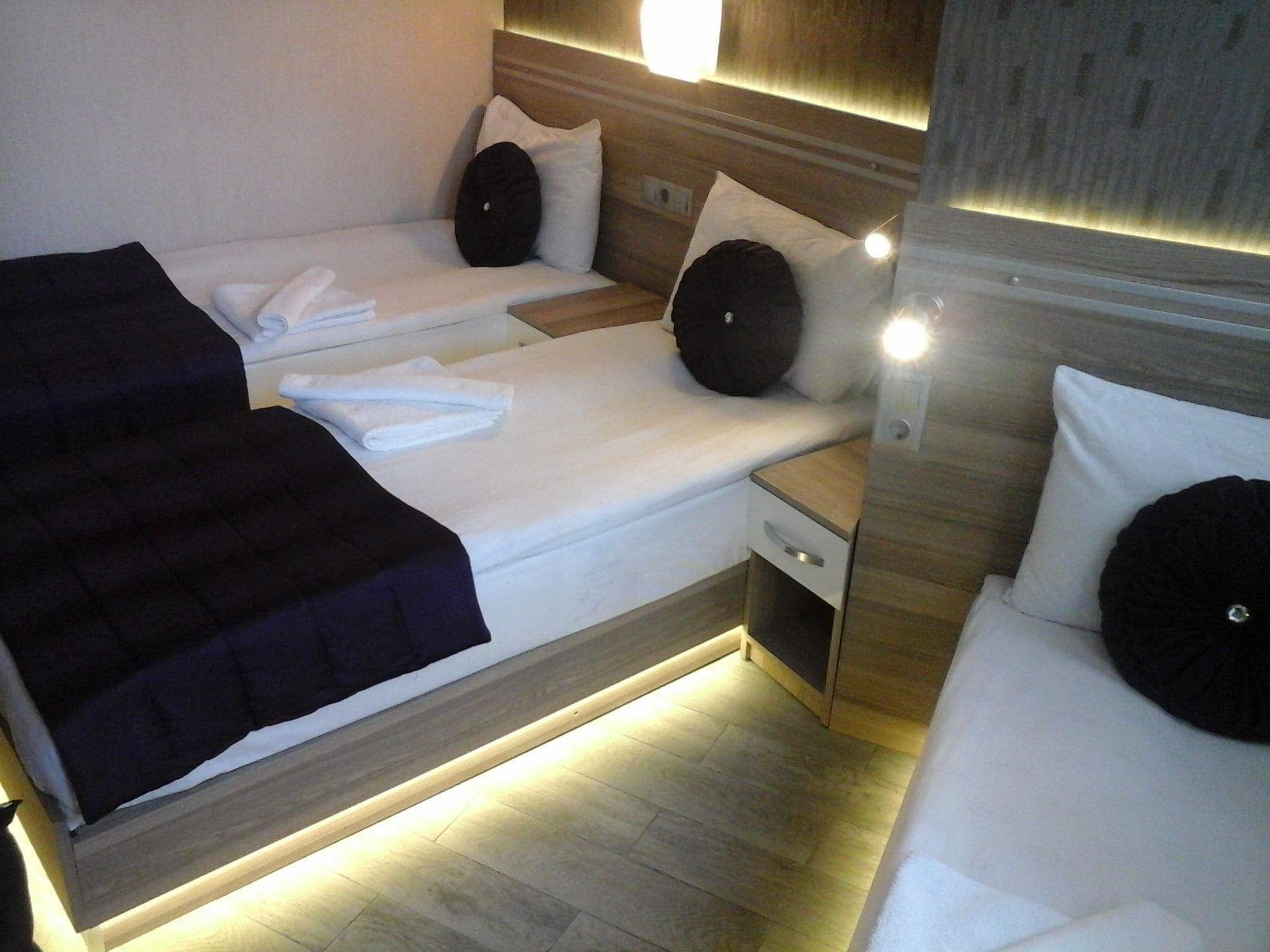 A compact hotel room with two beds, each with white linens and black pillows. There is a small bedside table with a drawer and a nightstand with a lamp.