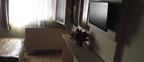 Una stanza piccola e scarsamente illuminata con un letto, un termosifone, una televisione e una finestra con tende.