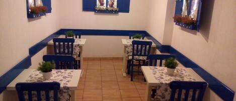 Un comedor con cuatro mesas y sillas, cada una con una planta en maceta. Las paredes tienen detalles azules y hay dos ventanas con contraventanas.