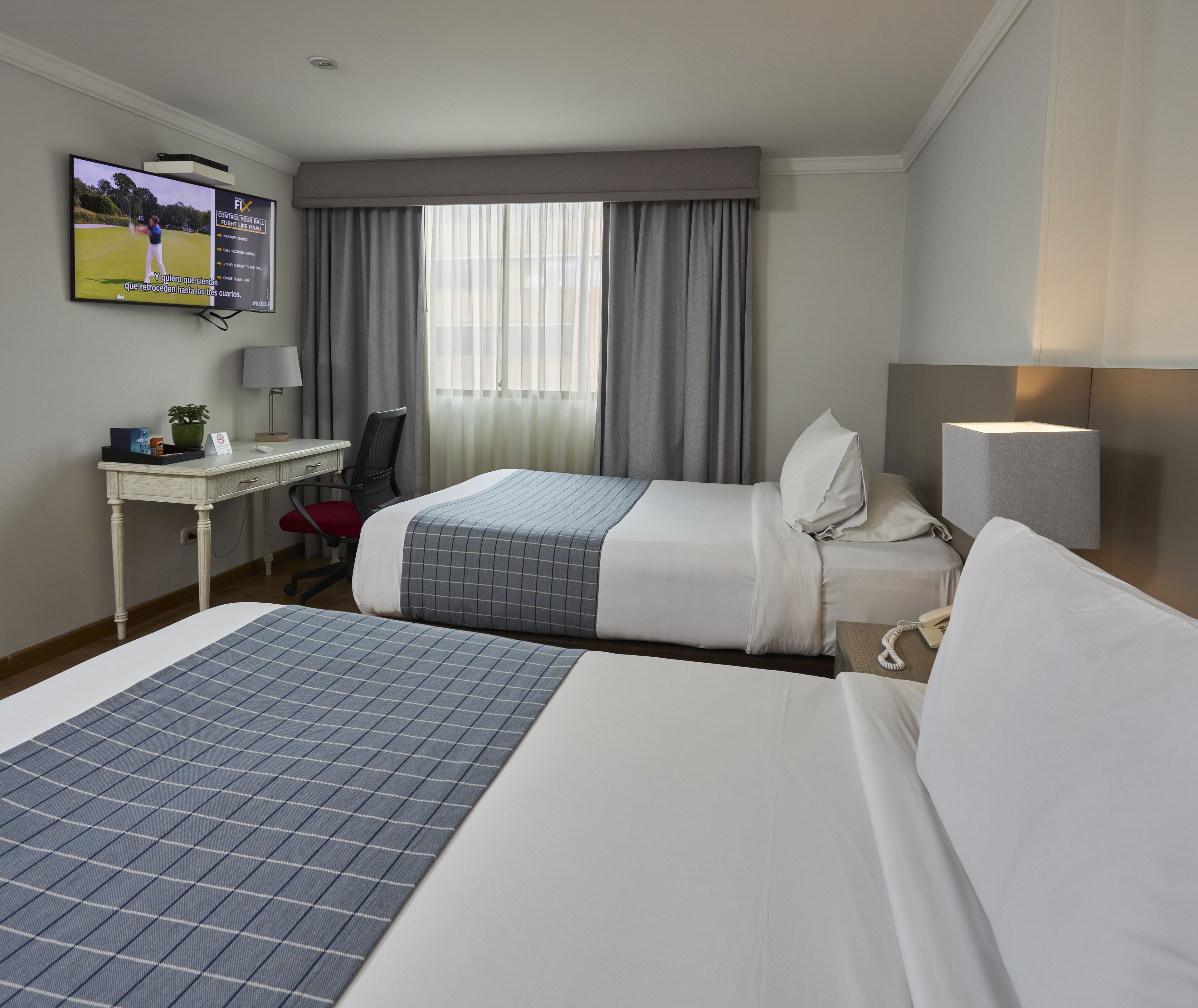 A hotel room with two beds, a desk, a TV, and a window with curtains.