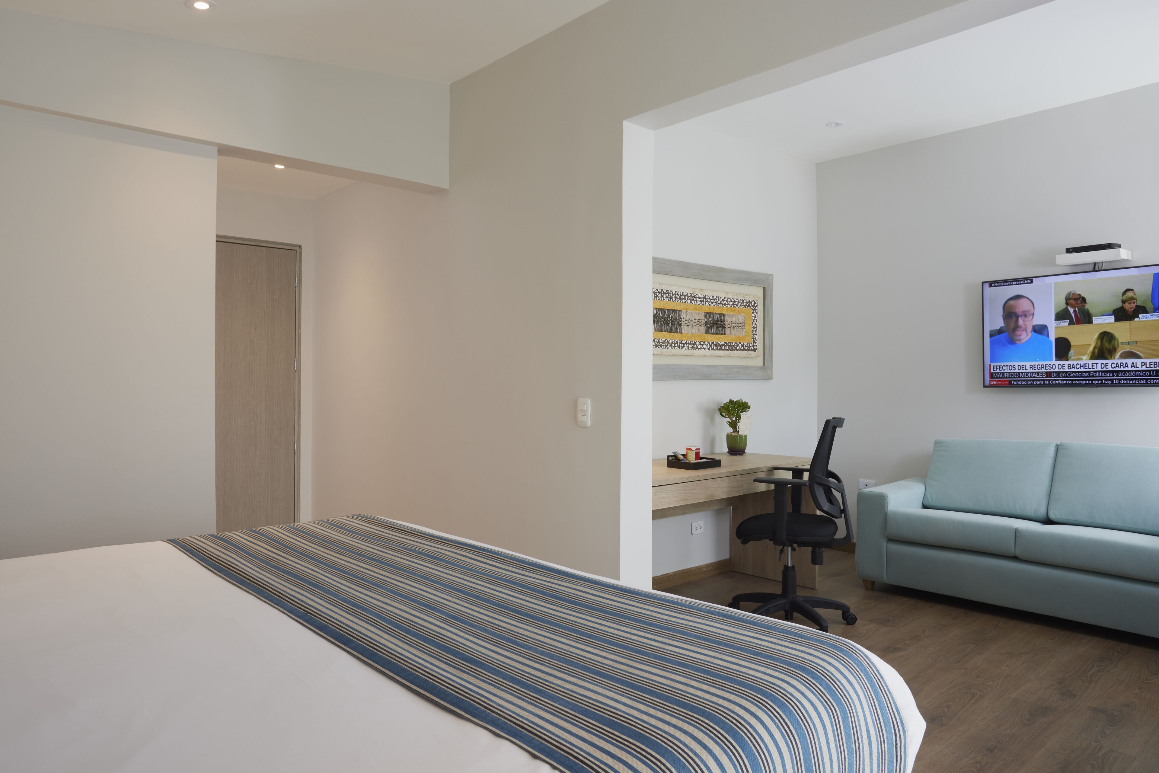 A hotel room with a bed, a desk with a computer, a couch, and a TV displaying a video conference.