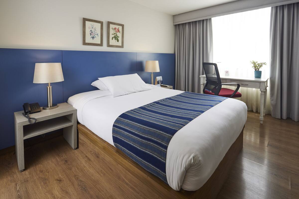 A hotel room with a bed, bedside lamps, a desk, a chair, and a window with curtains.