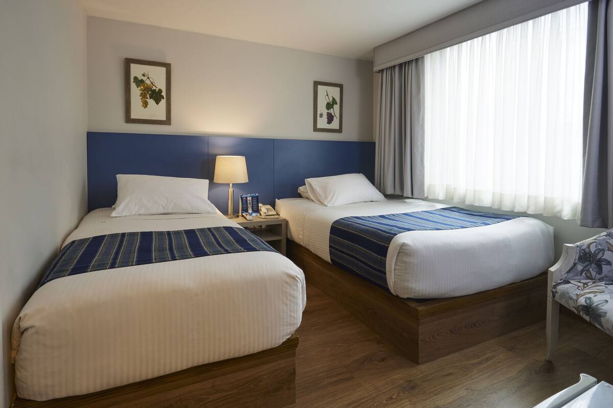 A hotel room with two beds, a nightstand with a lamp, and a chair.