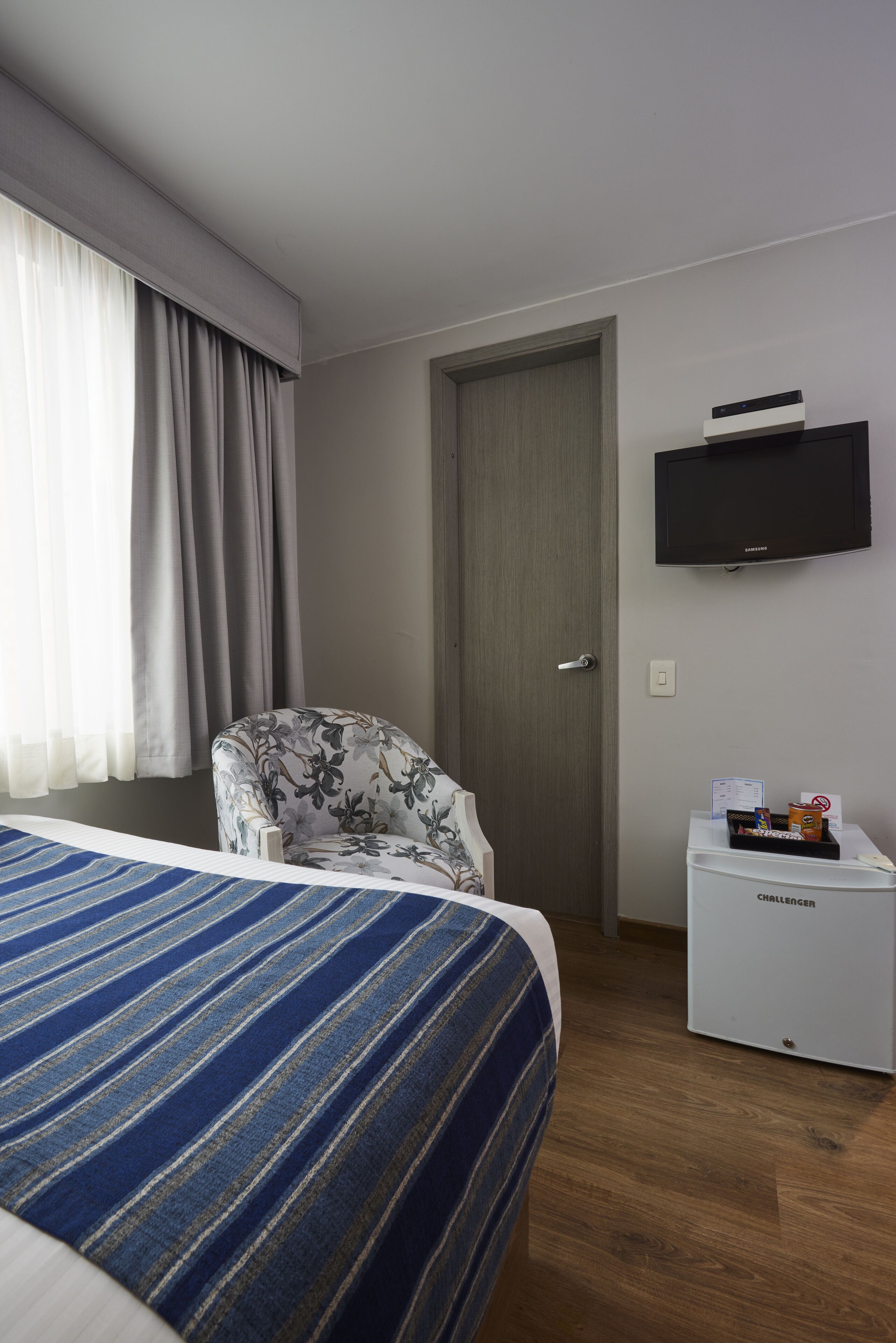A hotel room with a bed, a chair, a television, and a mini-fridge.