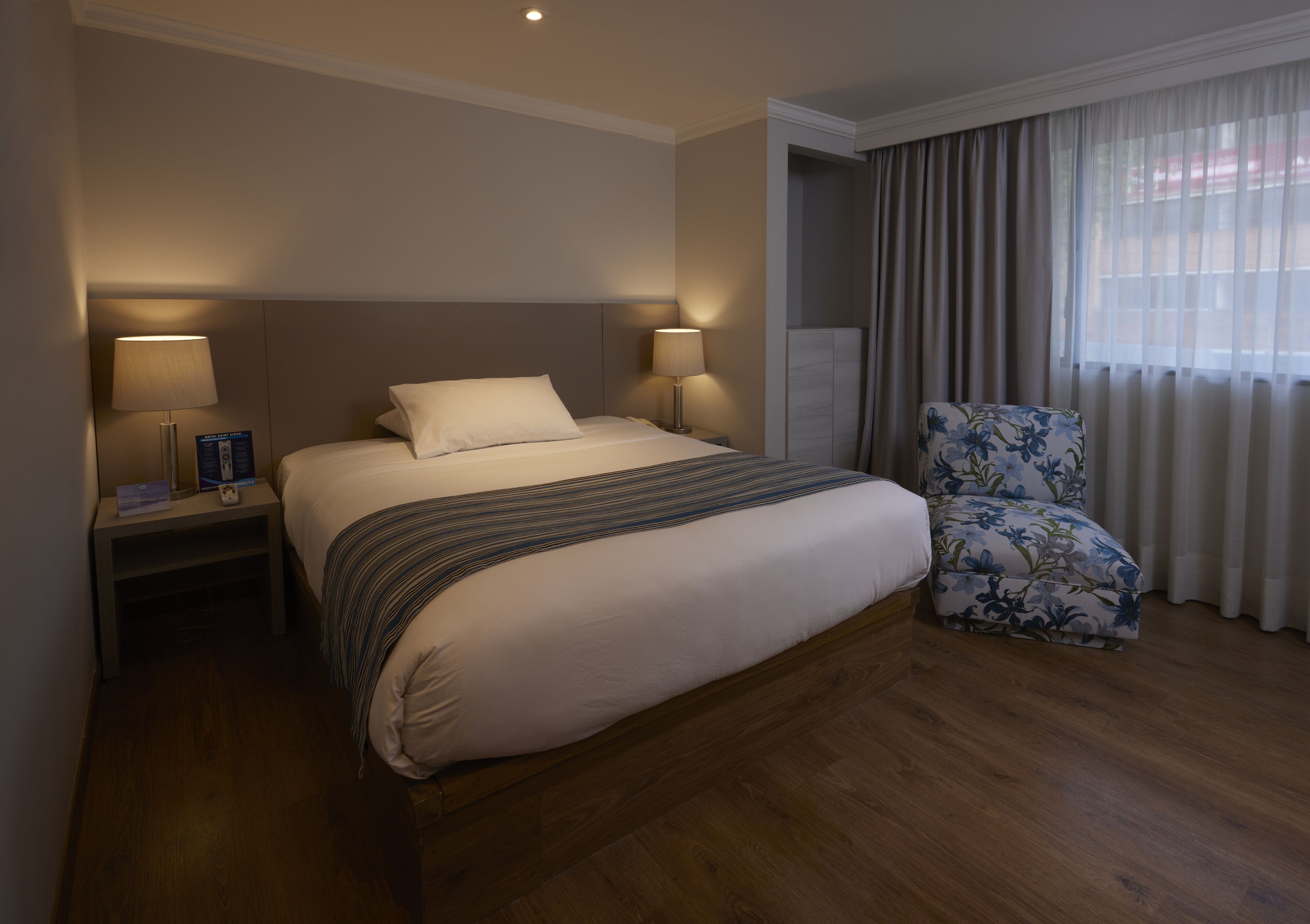 Ein Hotelzimmer mit einem großen Bett, zwei Nachttischlampen, einem blumigen Sessel und einem Fenster mit Vorhängen.