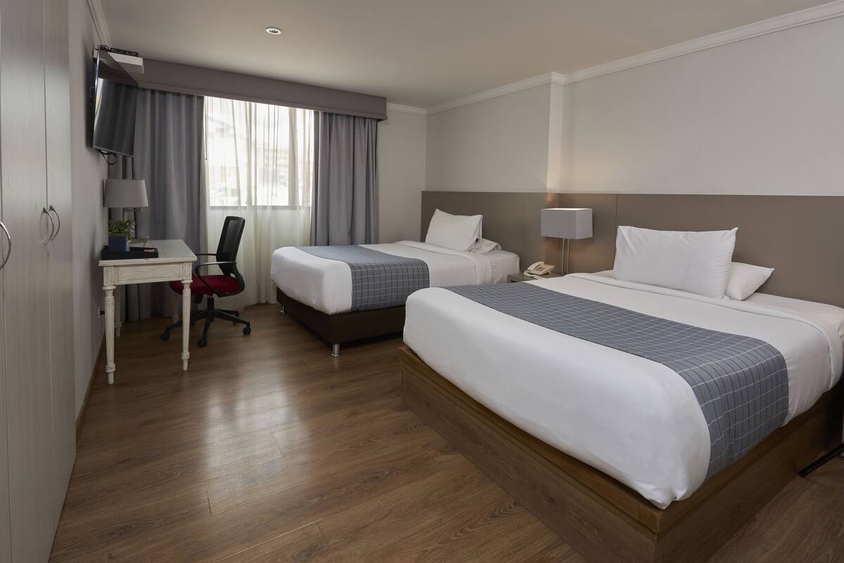 A hotel room with two beds, a desk with a chair, a TV, and a window with curtains.