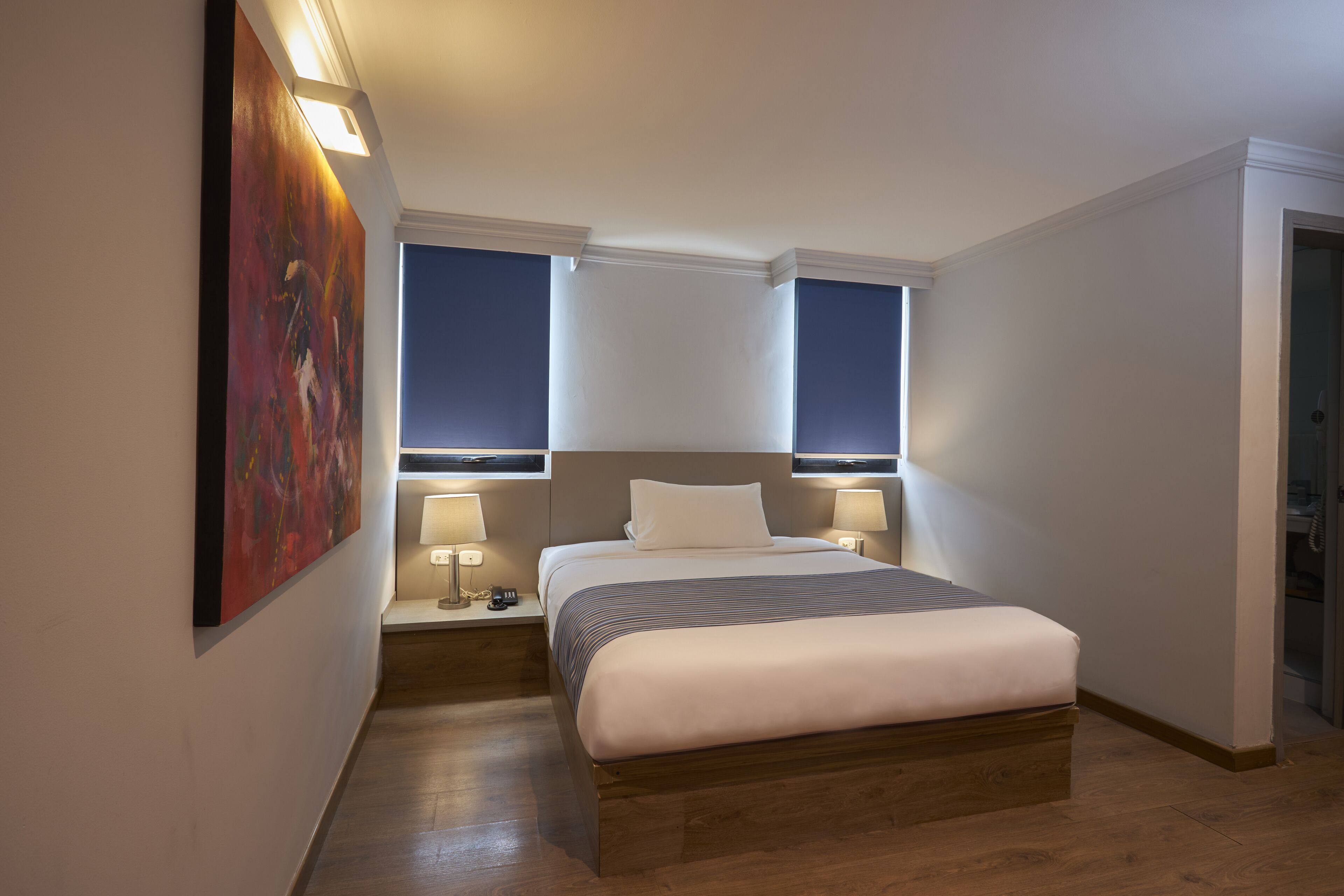 A modern hotel room with a large bed, two bedside tables with lamps, a painting on the wall, and a view of the night sky through the windows.