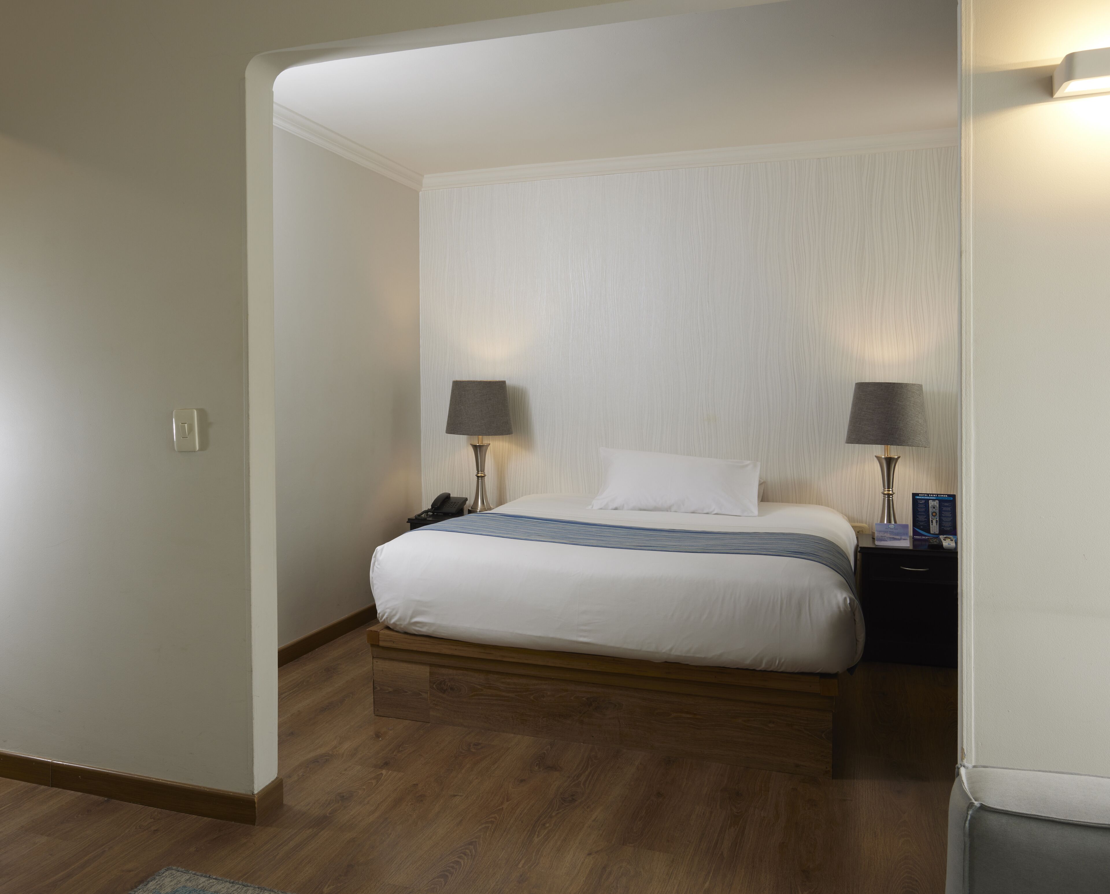 Ein Hotelzimmer mit Holzboden, einem Bett mit weißer Bettwäsche, zwei Nachttischlampen, einem Nachttisch und einer wandmontierten Leuchte.