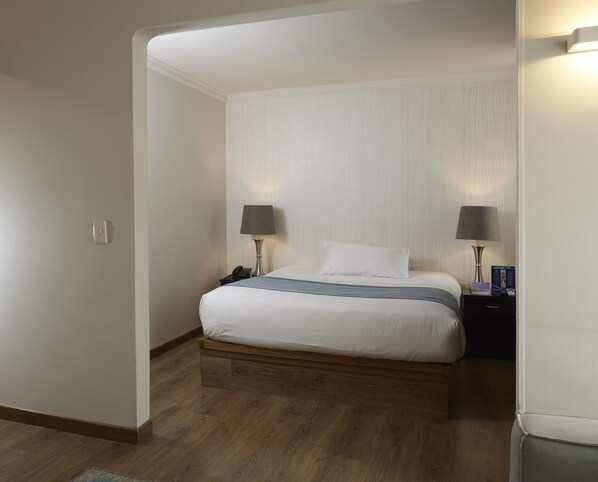 A hotel room with a wooden floor, a bed with white bedding, two bedside lamps, a nightstand, and a wall-mounted light fixture.