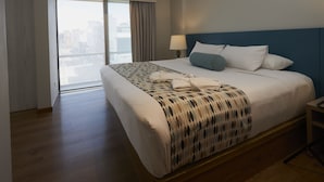 A hotel room with a large bed, a nightstand, a lamp, and a view of the city through a large window. - Hotel Saint Simon (Bogotá)