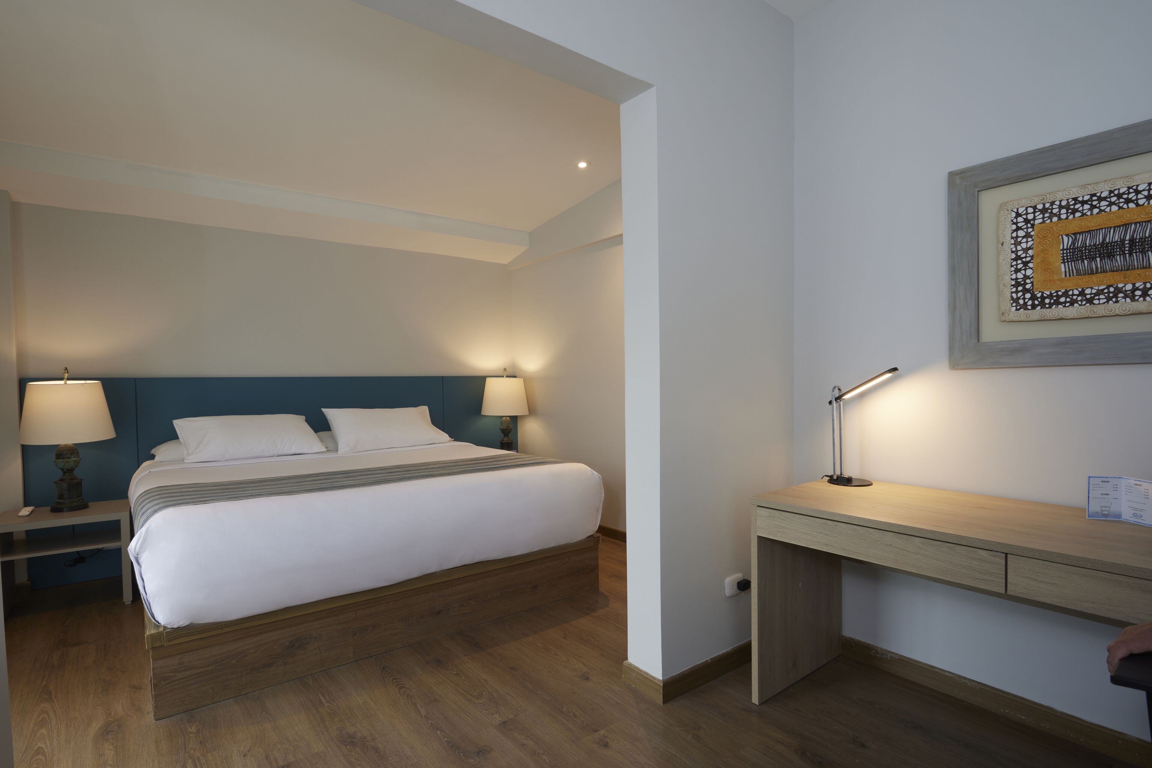 Una habitación de hotel con una cama, lámparas de noche, un escritorio y una obra de arte enmarcada en la pared.