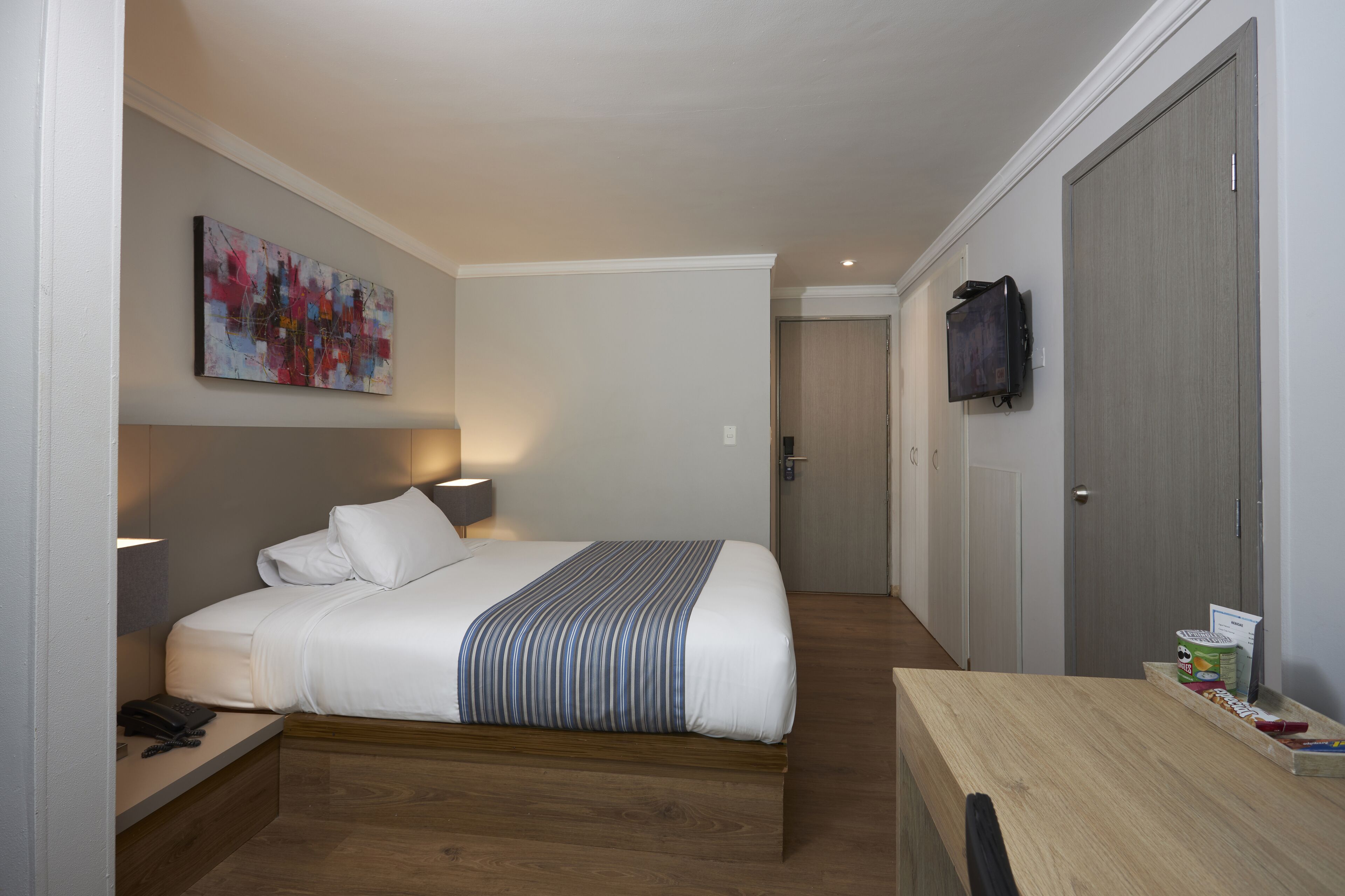 Hotelkamer met een bed, nachtkastje, lamp, kunstwerk, televisie en een bureau met spullen erop.