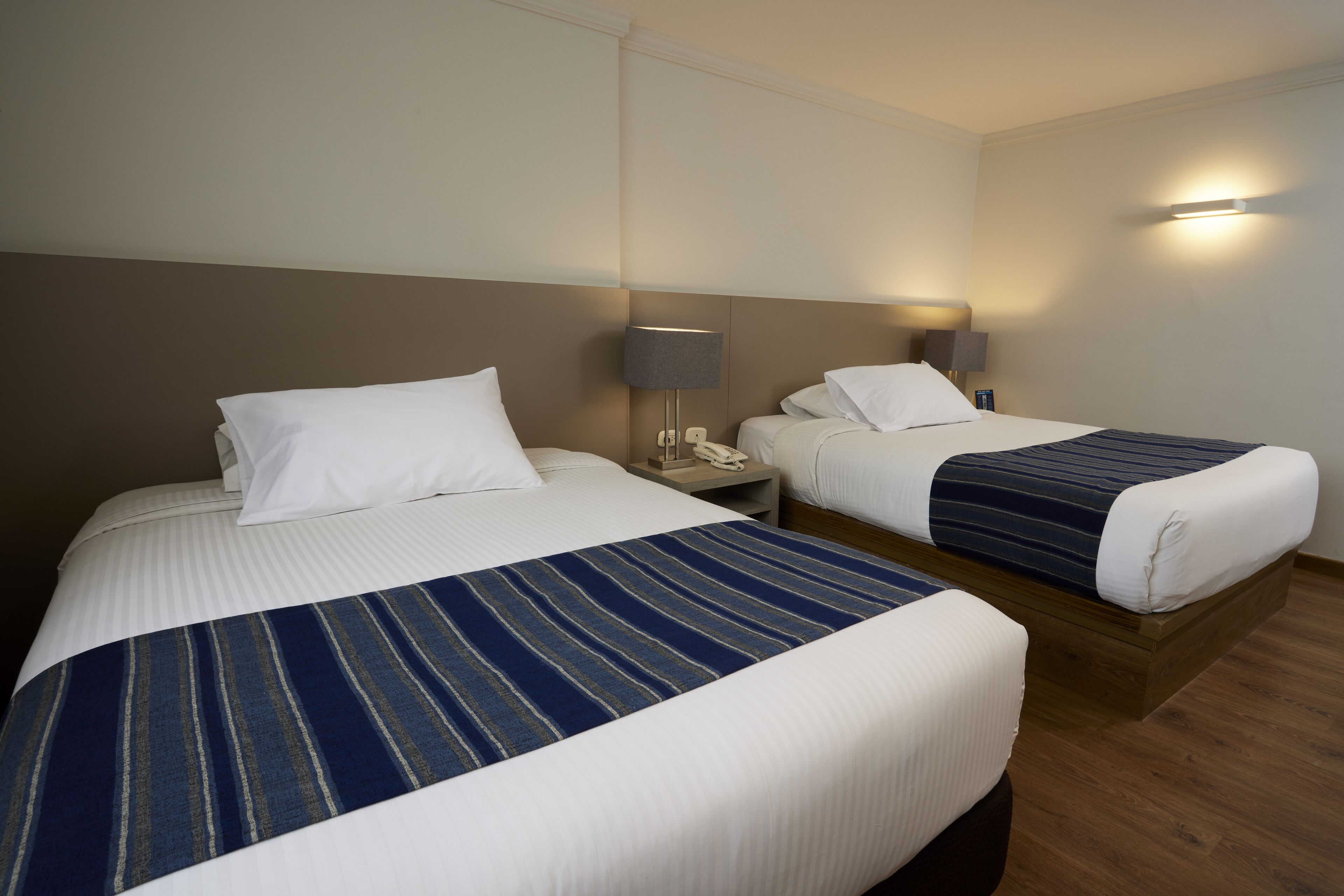 Ein Hotelzimmer mit zwei Betten, jeweils mit weißer Bettwäsche und einer blau gestreiften Decke. An jedem Bett befindet sich ein Nachttisch mit einer Lampe und einem Telefon.