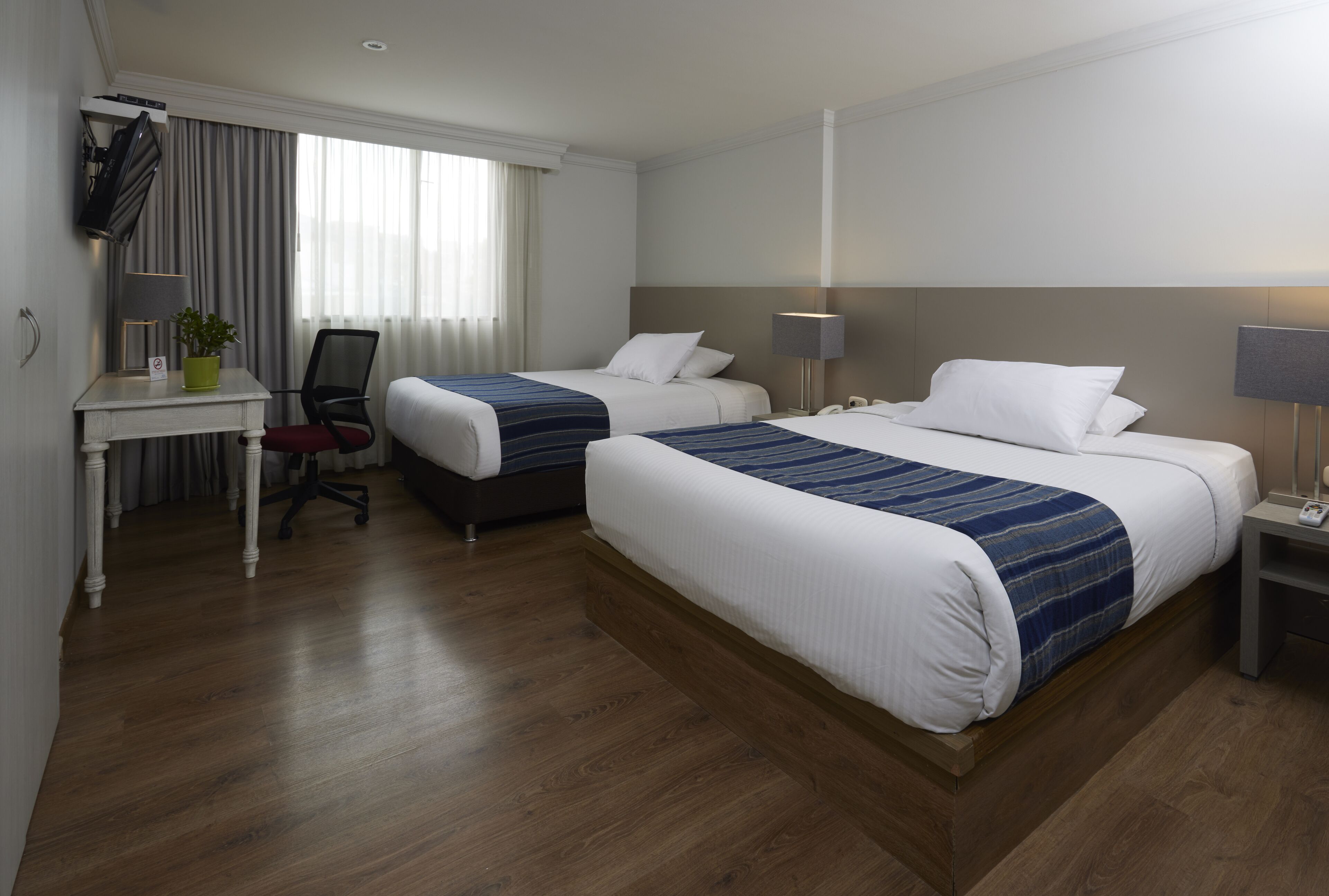 A hotel room with two beds, a desk with a chair, a TV, and a lamp.