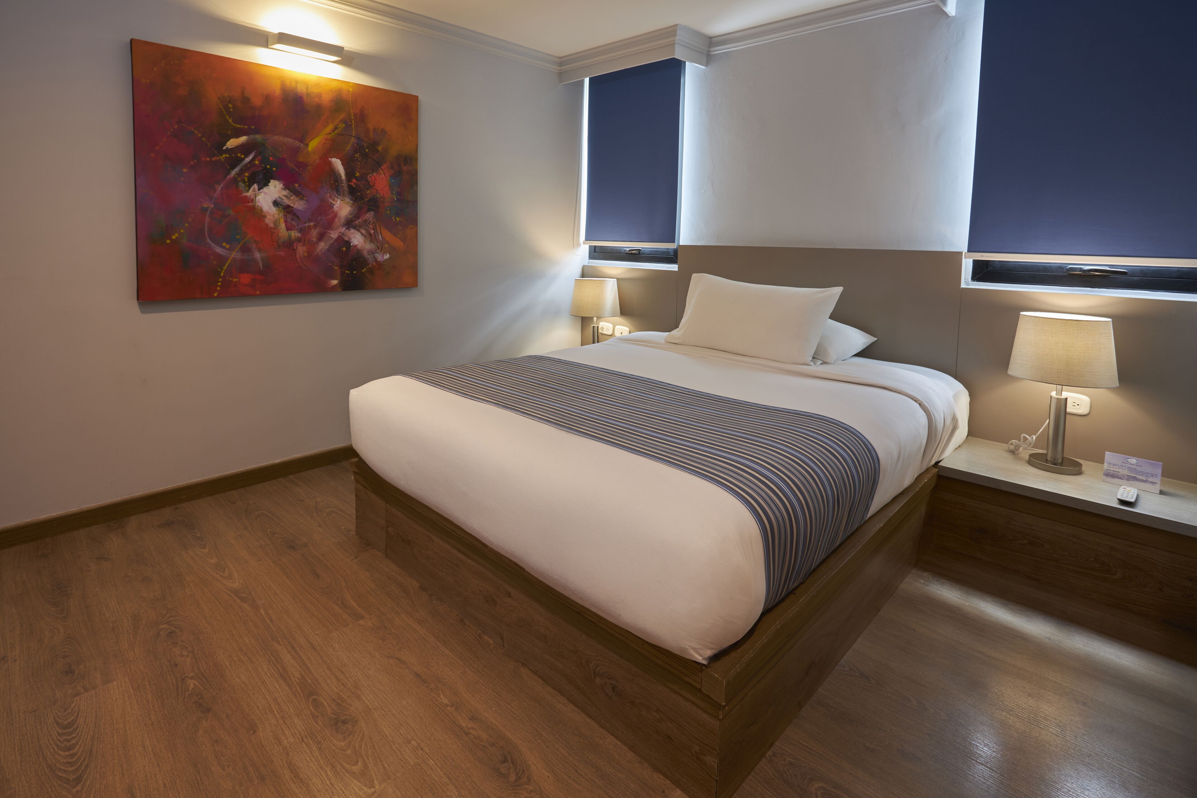 Une chambre d’hôtel avec un grand lit, deux tables de chevet avec des lampes, un tableau au mur et une fenêtre avec des stores.