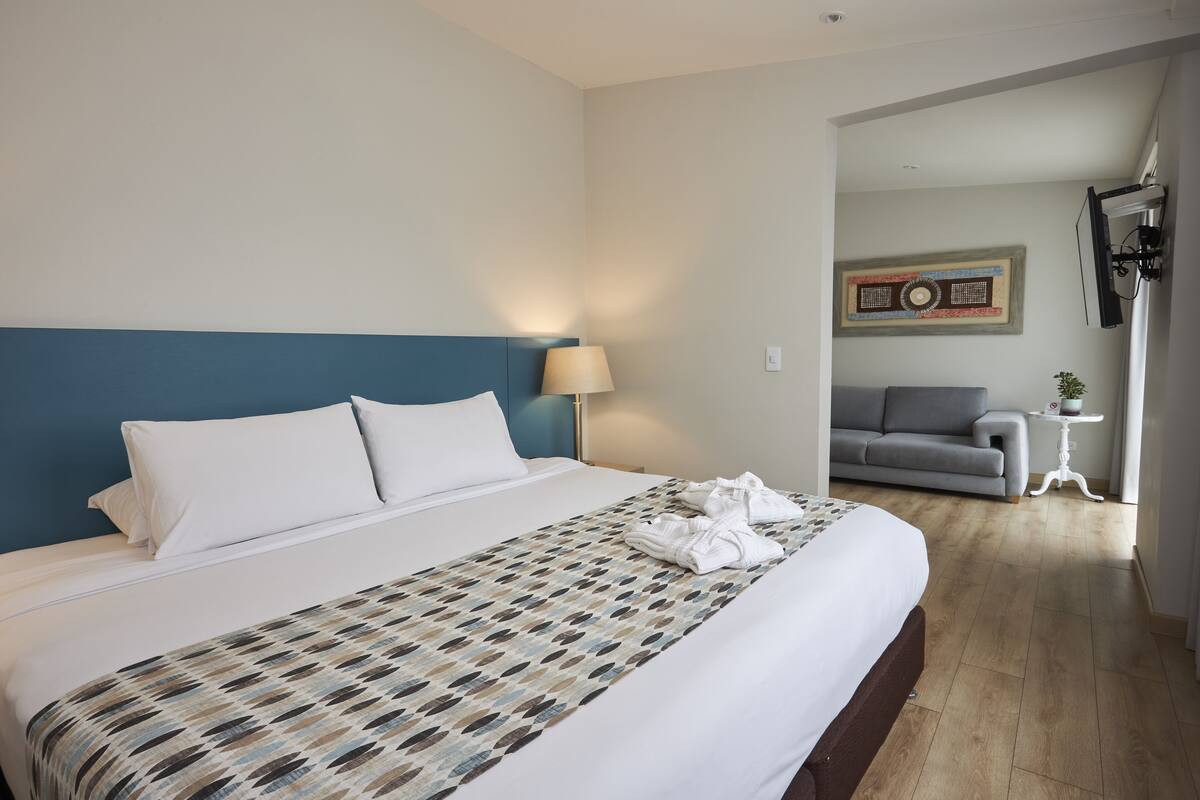A hotel room with a large bed, a blue headboard, a patterned bedspread, and a grey sofa in the background.