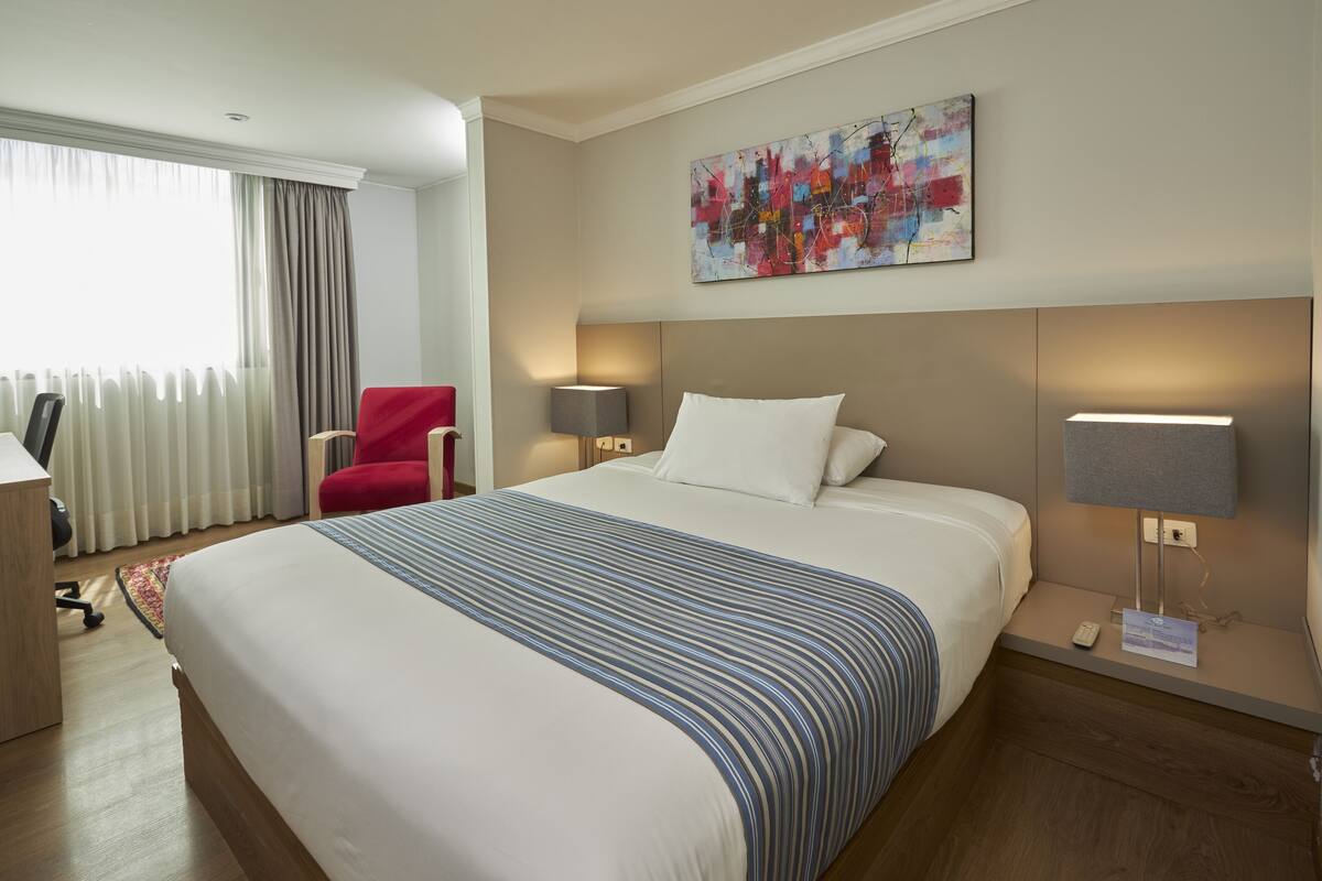 A hotel room with a large bed, a red chair, a desk, and a window with curtains.