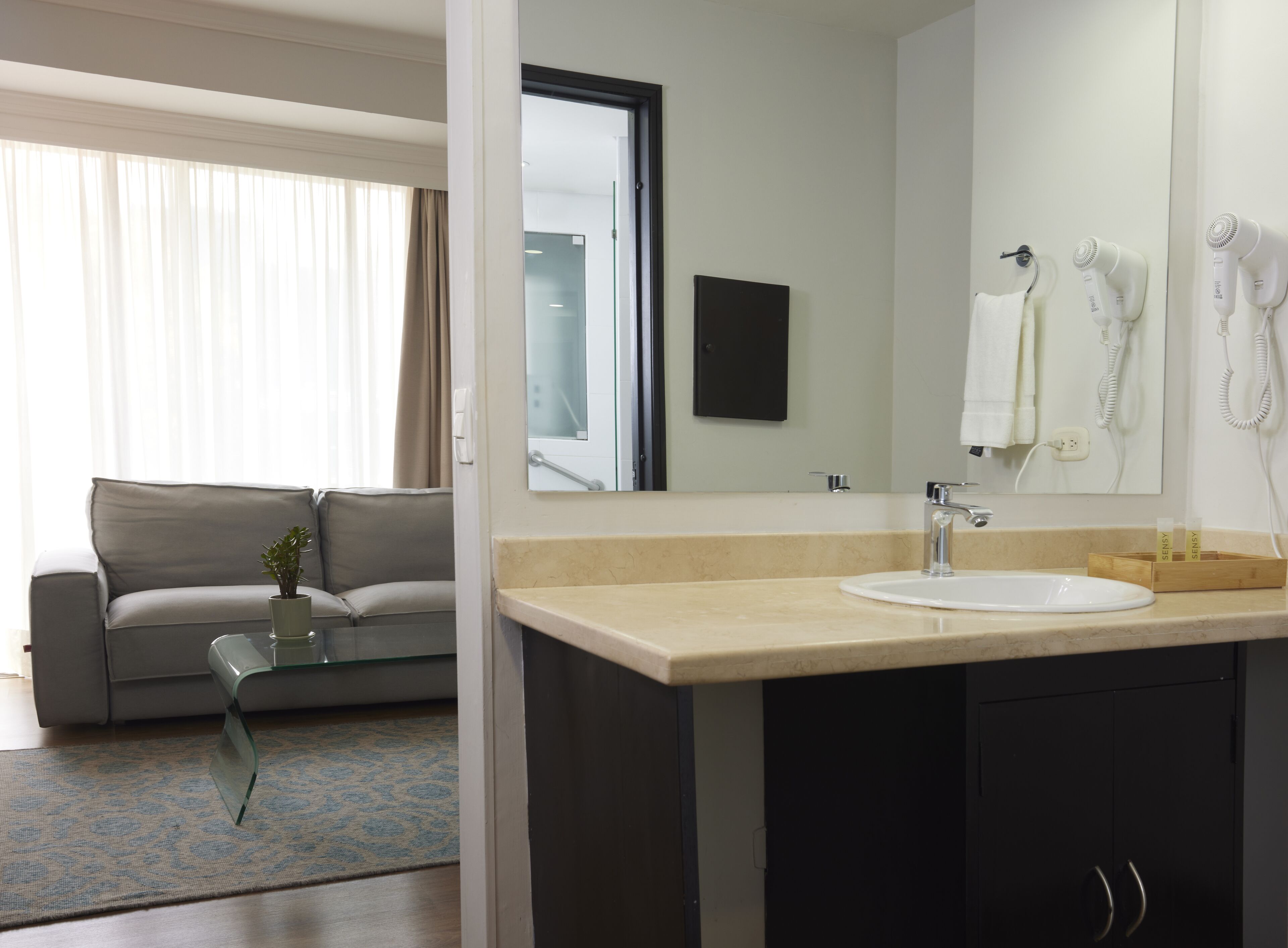 A hotel room with a sofa, a glass coffee table, a sink, and a mirror.